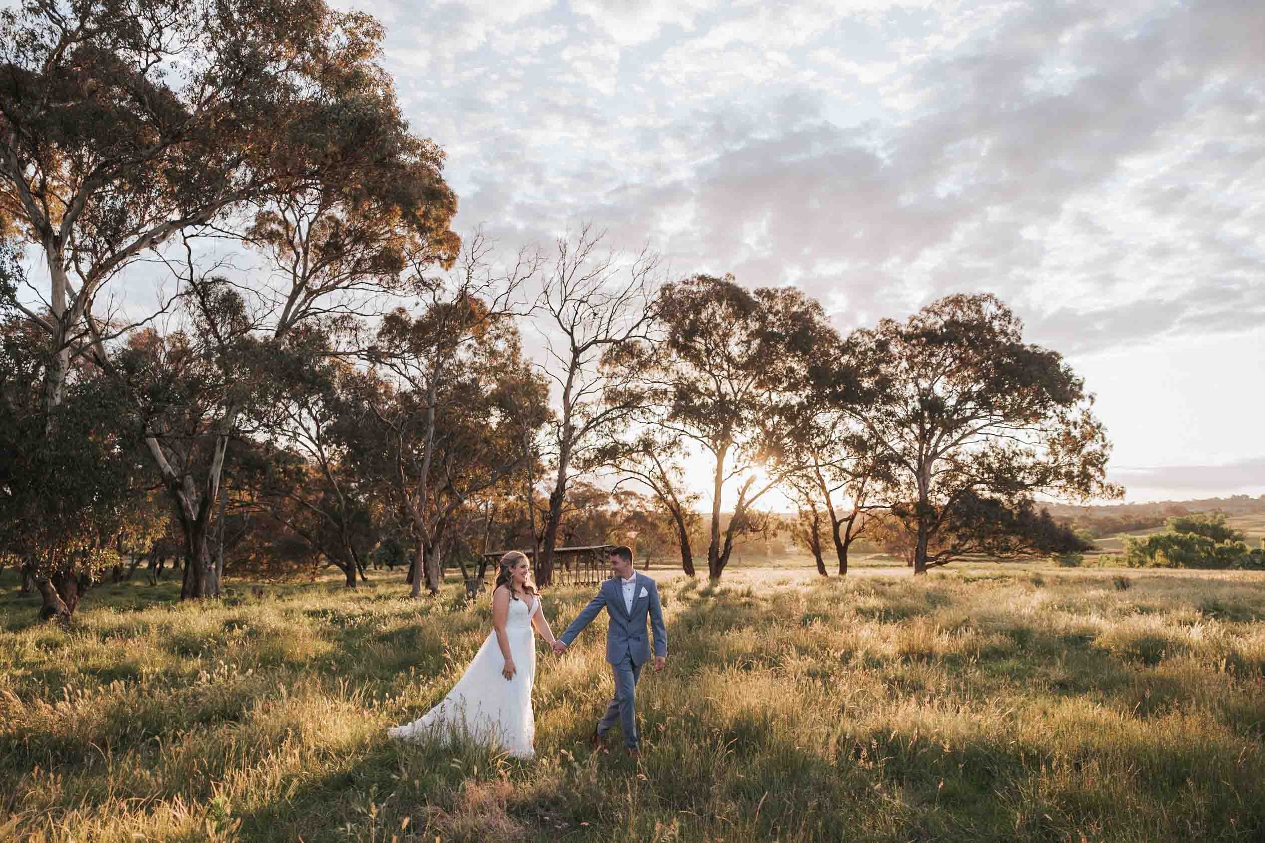 A bride and groom walking hand in hand through a grassy field at sunset, with trees and a cloudy sky in the background.