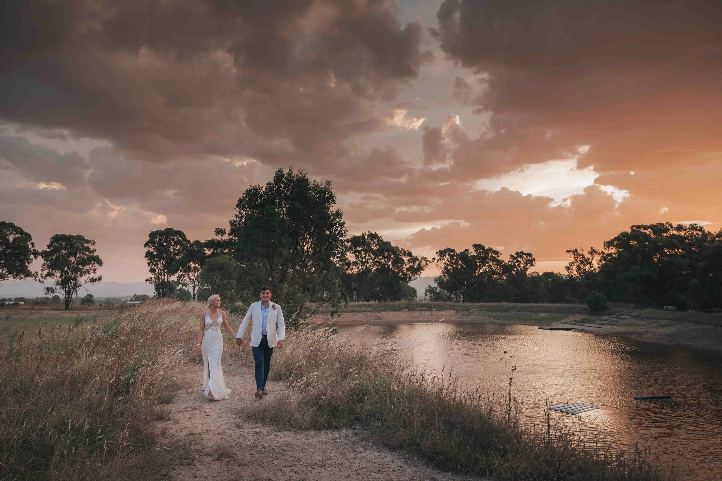 A newlywed couple walking hand-in-hand along a dirt path next to a body of water at sunset. The bride is wearing a white wedding dress, and the groom is in a light-colored blazer and dark pants. Clouds are overhead with the sky glowing in warm colors