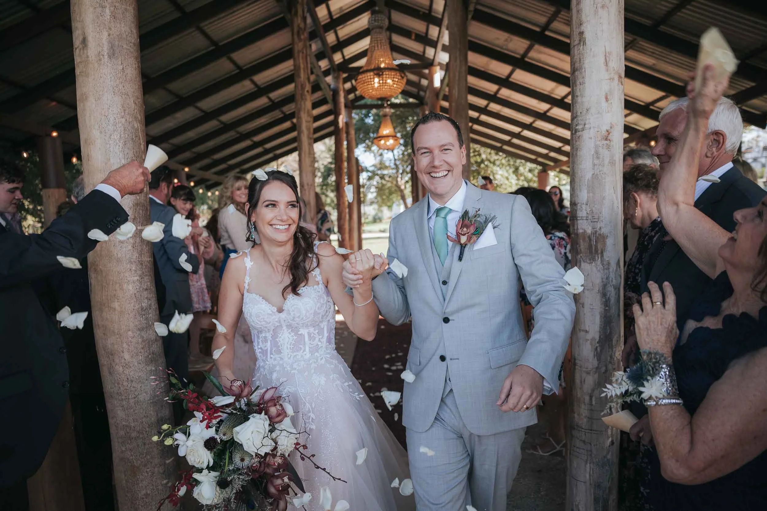 A bride and groom smiling and holding hands as they walk through a shower of flower petals, surrounded by wedding guests in an open-air wooden pavilion.