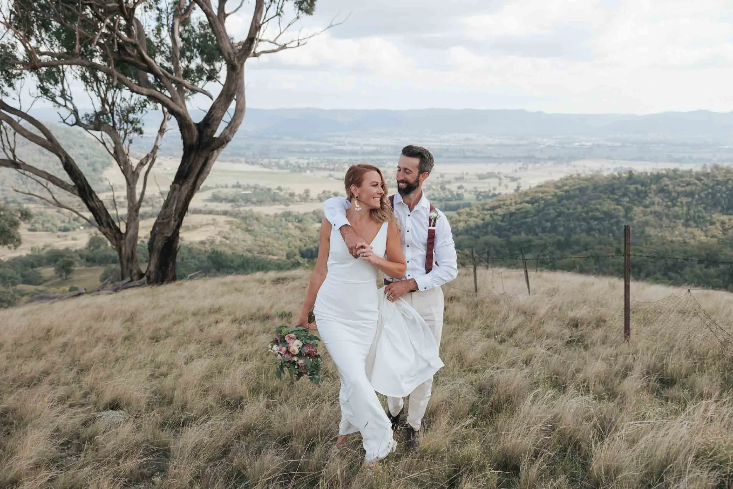 A couple walking together in a grassy field, smiling and looking at each other, with a scenic landscape of hills and trees in the background.
