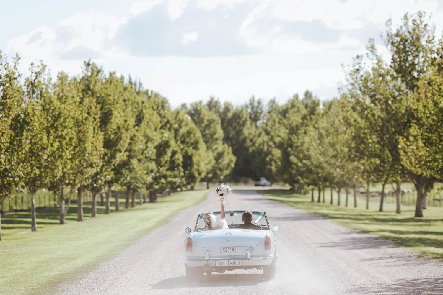A vintage convertible car driving down a tree-lined country road with a bride and groom inside, the bride holding a bouquet and raising her arm in celebration.