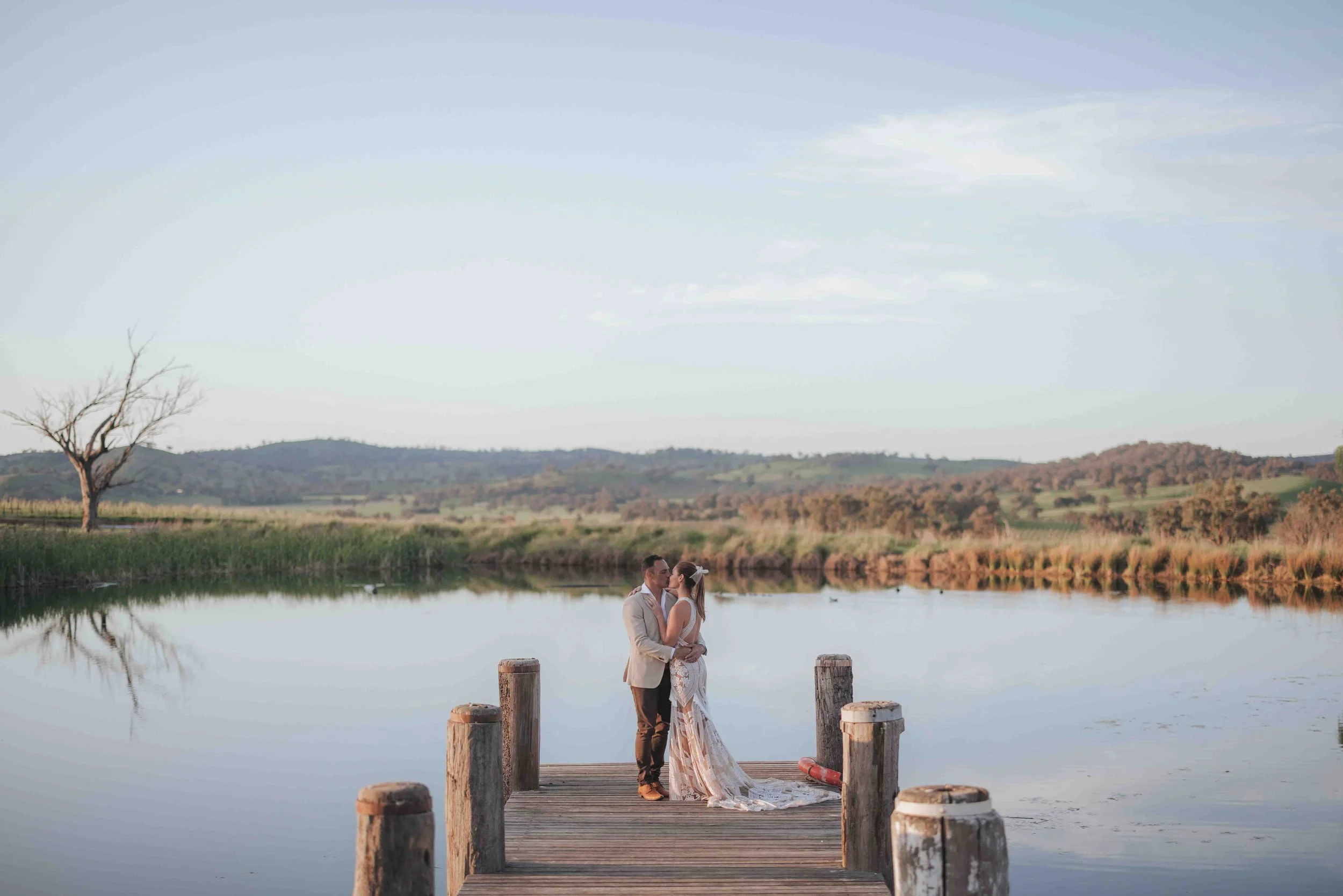 A newlywed couple stands on a wooden dock by a tranquil lake, sharing an intimate moment with a scenic countryside in the background.
