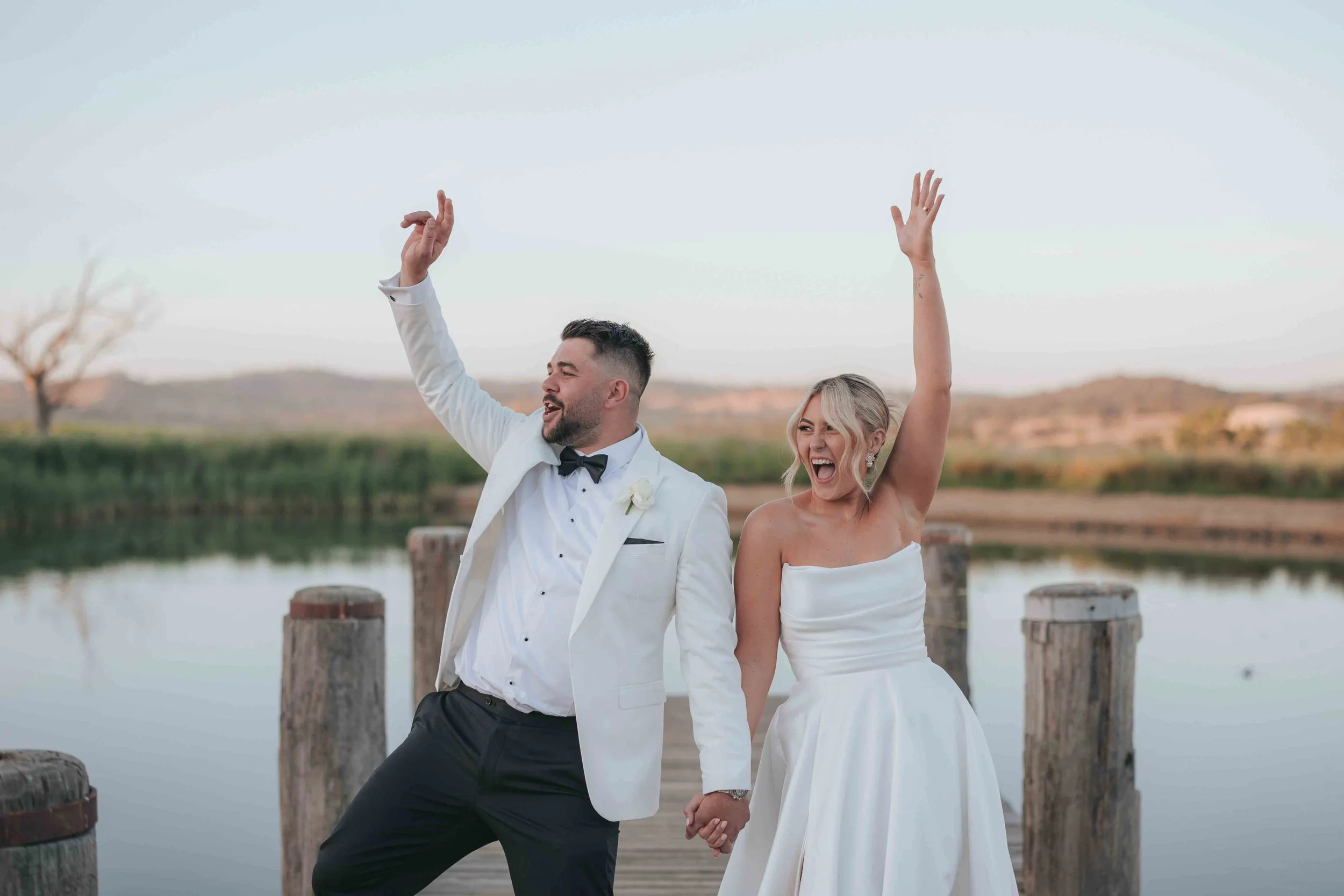 A happy bride and groom in wedding attire on a dock by a lake, holding hands, dancing, and celebrating outdoors during sunset.