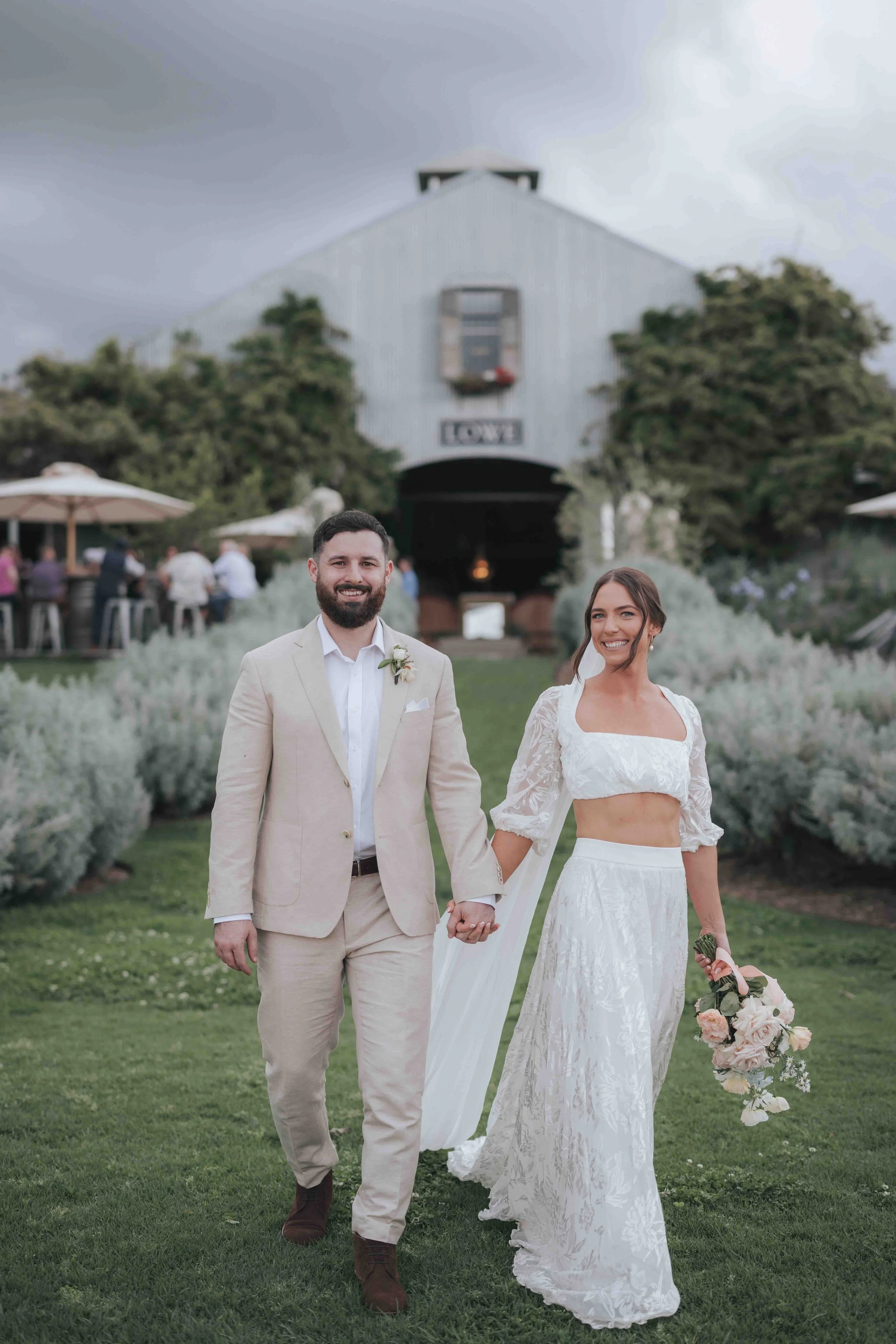 A newlywed couple walking hand in hand outside at a rustic wedding venue, smiling, with a barn and guests in the background.