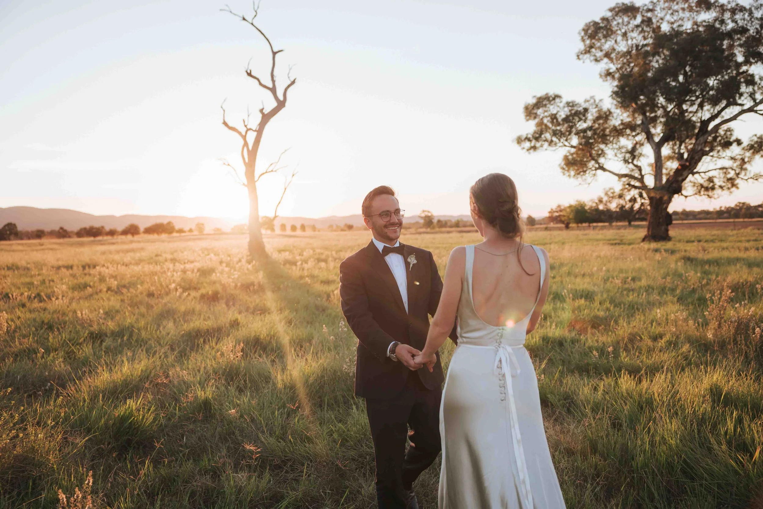 A bride and groom holding hands and smiling at each other in a field at sunset with trees and distant hills in the background.