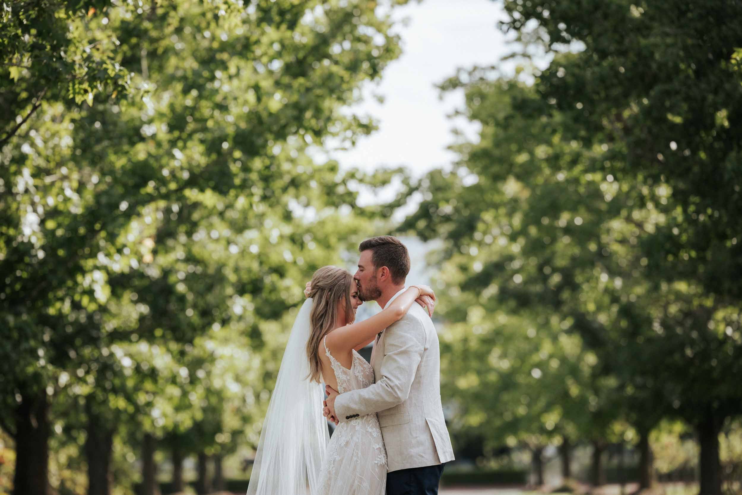 A bride and groom embrace outdoors during wedding, trees in background, bride wearing a lace wedding gown and veil, groom in a light-colored suit jacket.
