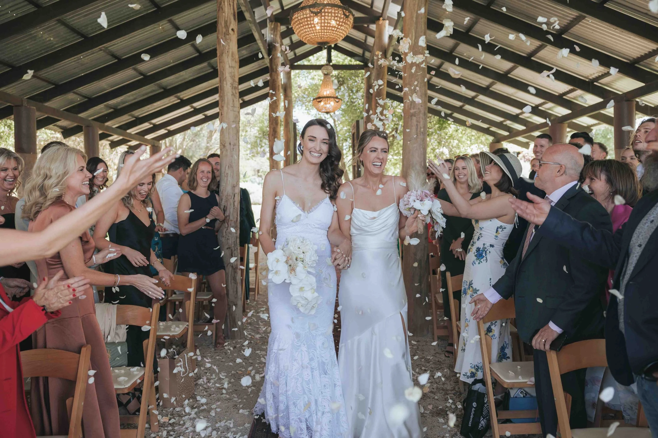 Two women in white wedding dresses walking down the aisle at a wedding reception, surrounded by smiling guests tossing flower petals, under a wooden pavilion with greenery outside.