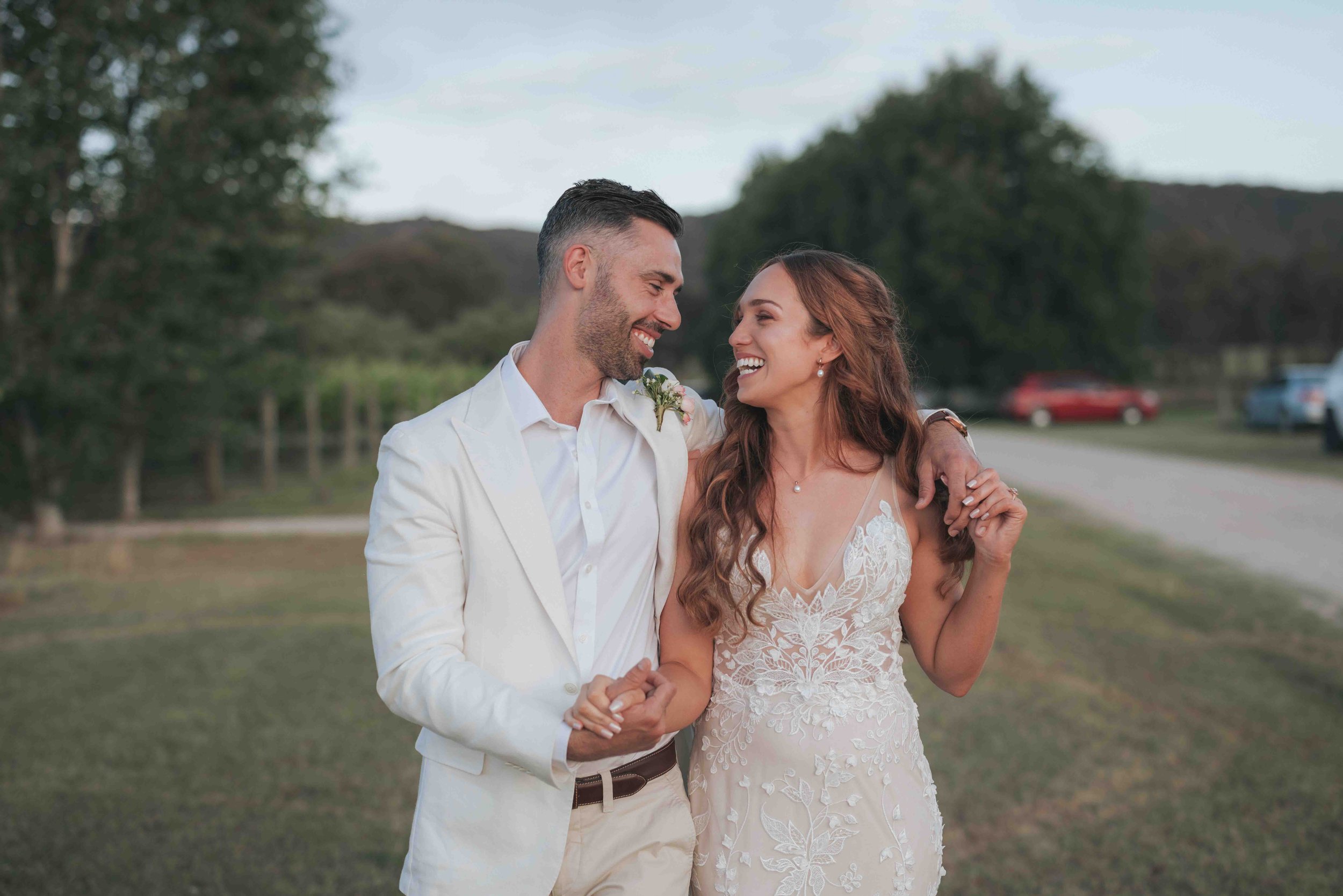 A happy couple dressed in wedding attire, standing outdoors on a grassy area with trees and parked cars in the background, smiling and holding hands.