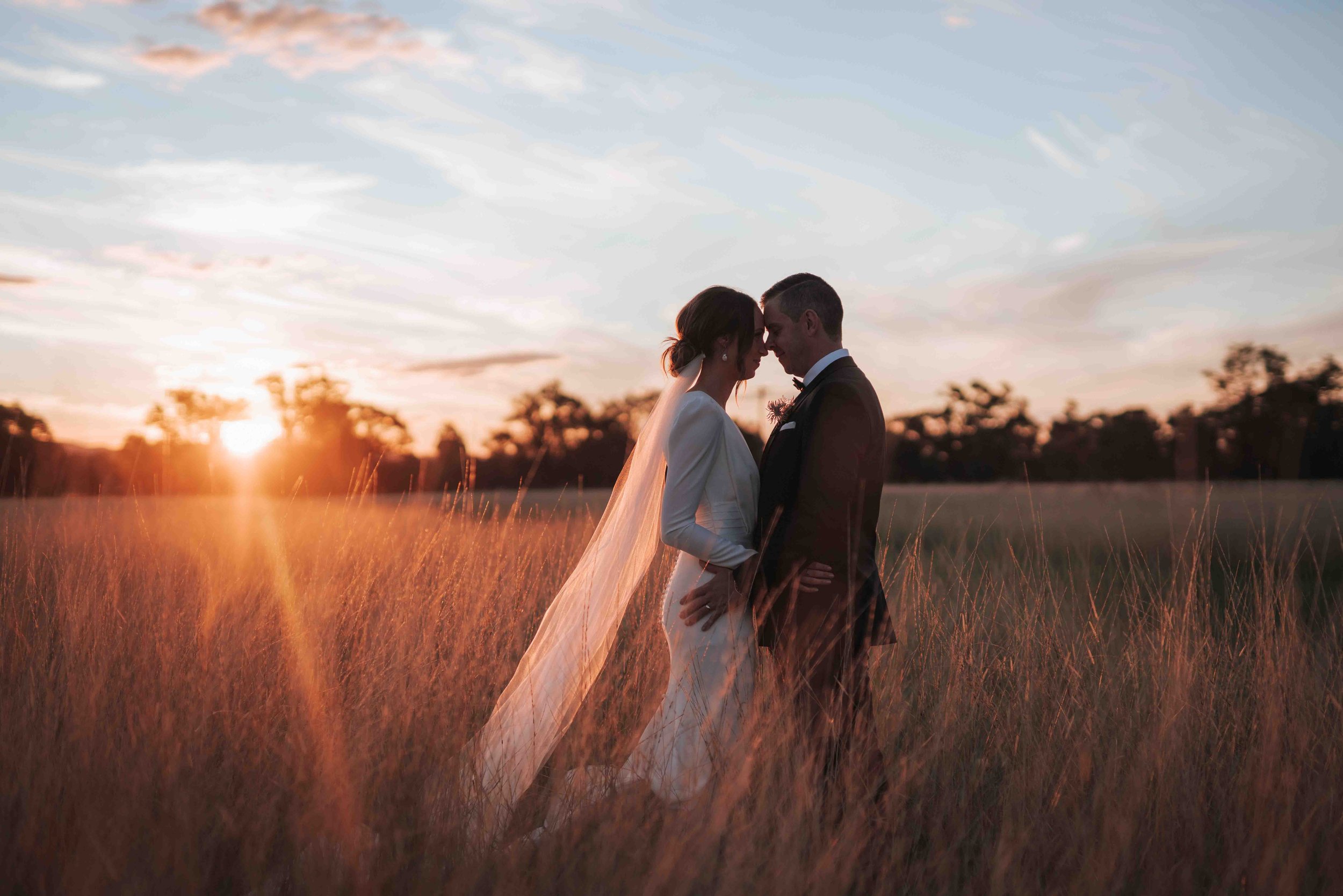 A bride and groom standing close together in a field at sunset, with their foreheads touching and holding each other, during their wedding.
