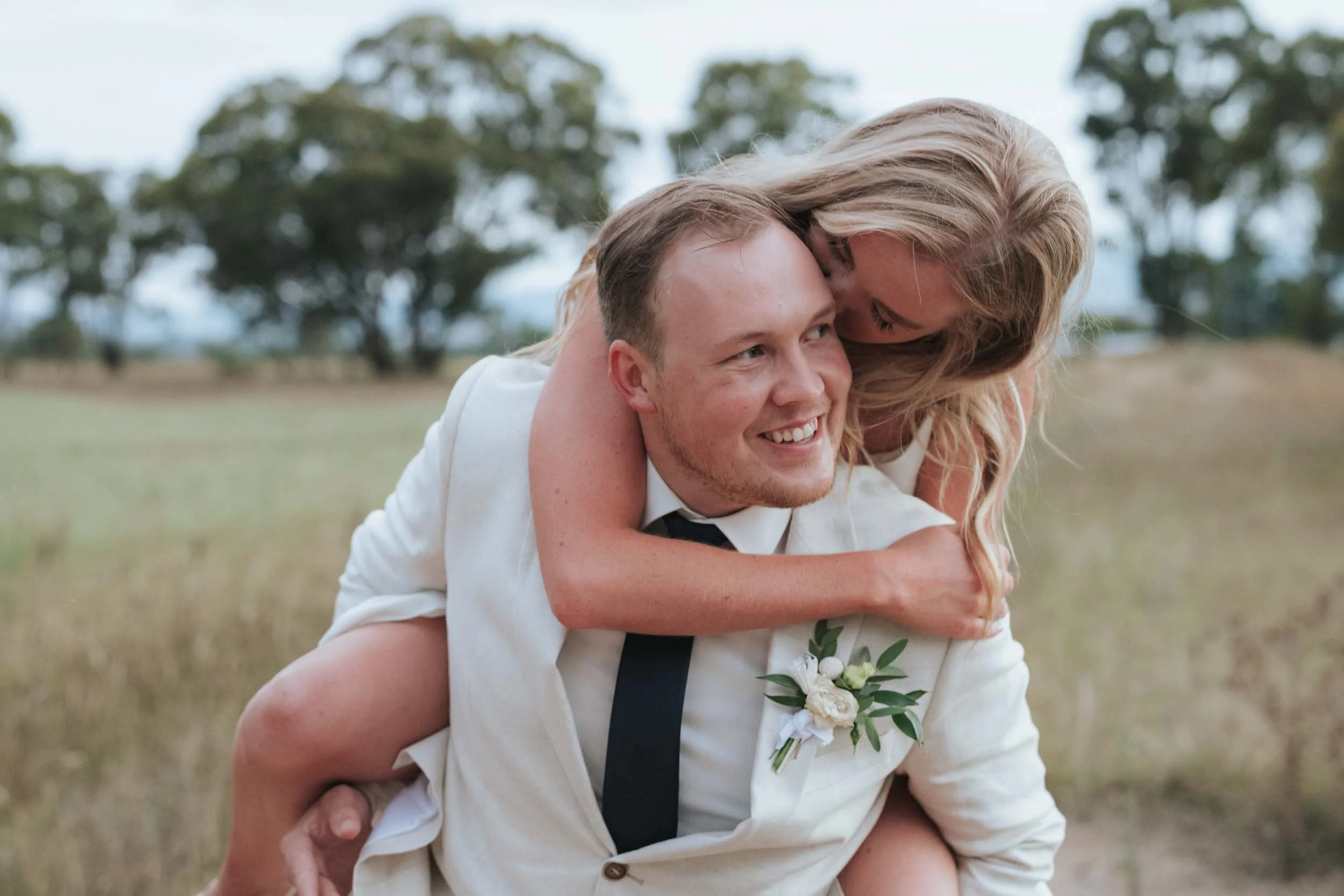 A happy couple outdoors; the woman is giving the man a piggyback ride and they are smiling at each other. The background features trees and a grassy field.