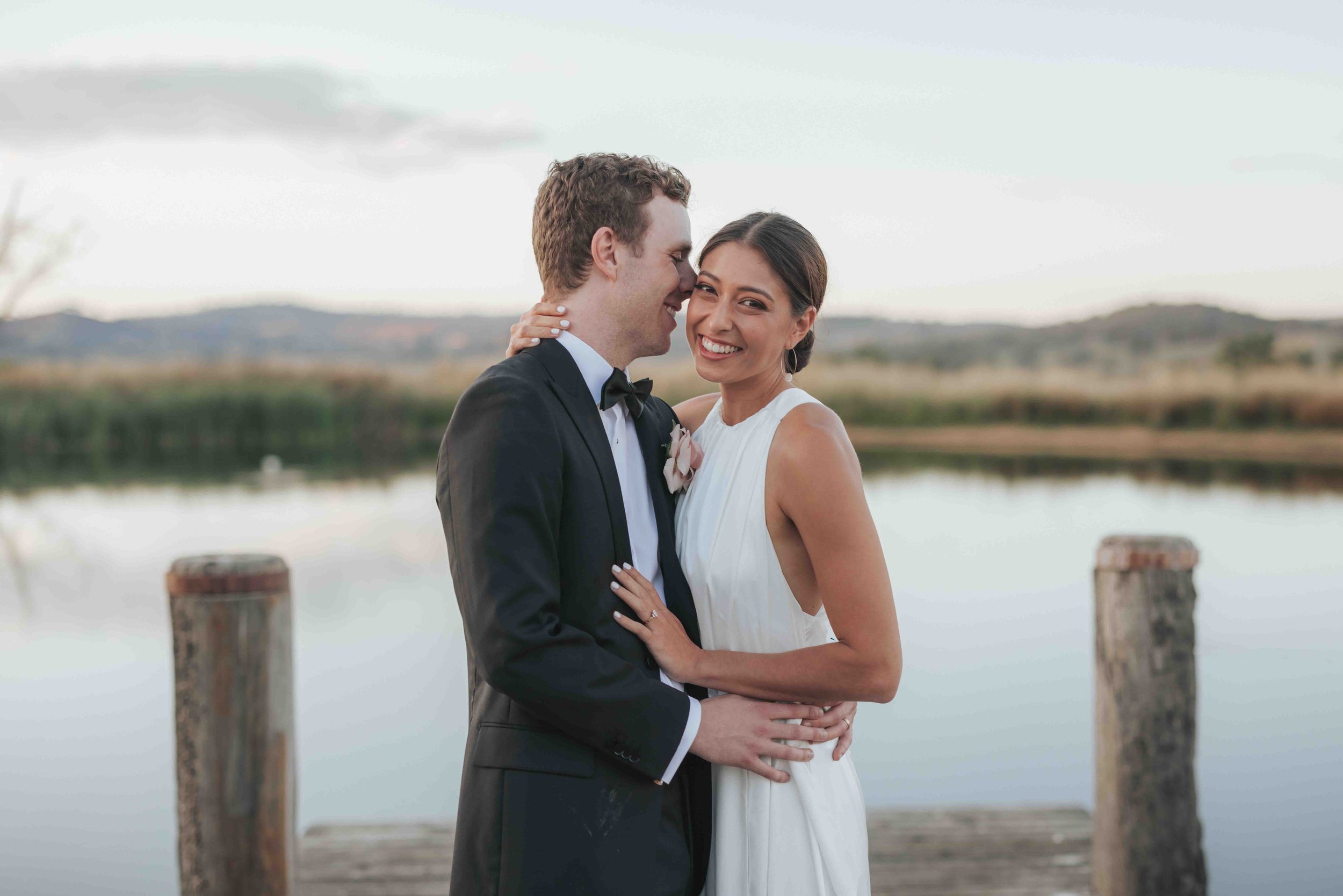 A newlywed couple embracing on a dock by a lake during sunset, smiling at the camera with the scenic landscape in the background.