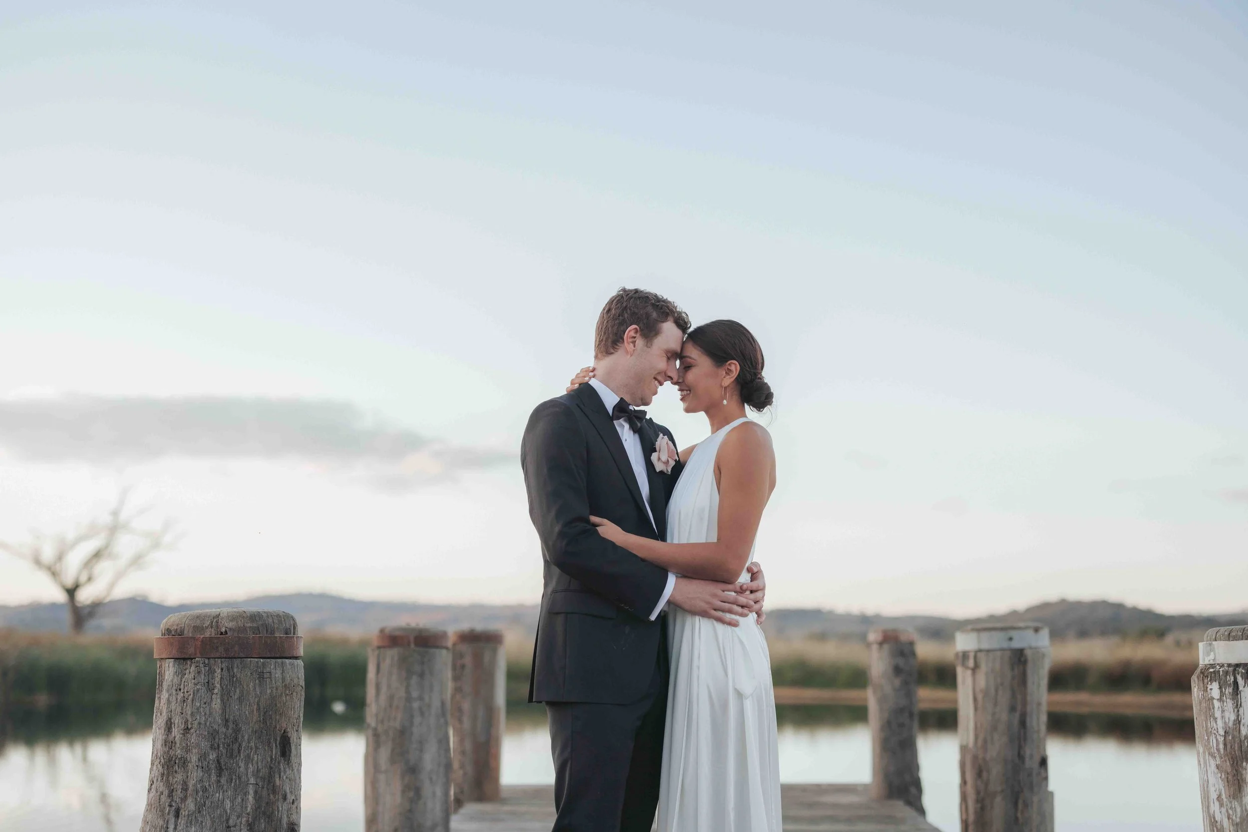 A newlywed couple, a man in a tuxedo and a woman in a white wedding dress, embrace on a wooden dock by a lake with a scenic landscape and a cloudy sky in the background.