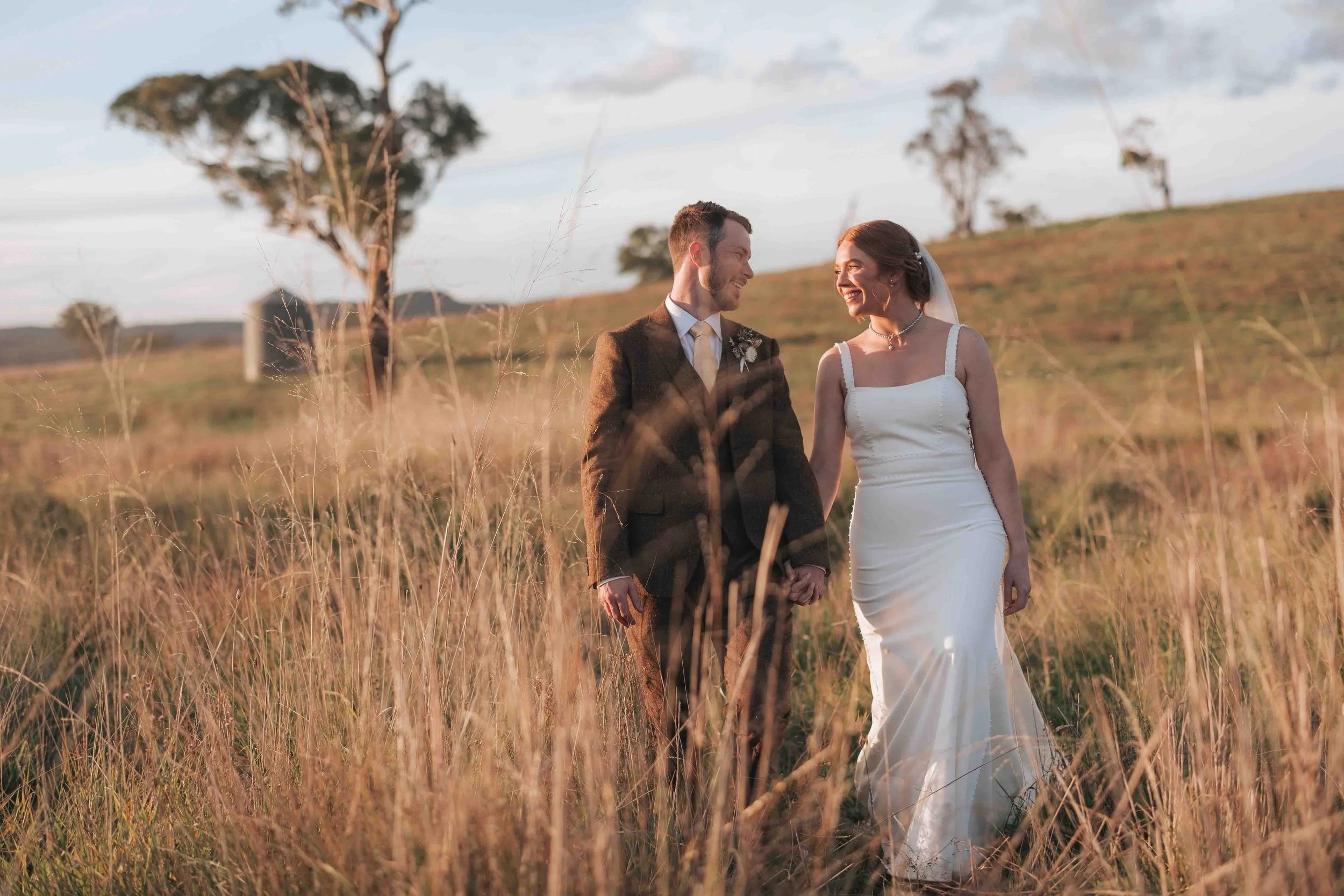 A newlywed couple holding hands and smiling at each other in a grassy field during sunset, with trees and a hilly landscape in the background.
