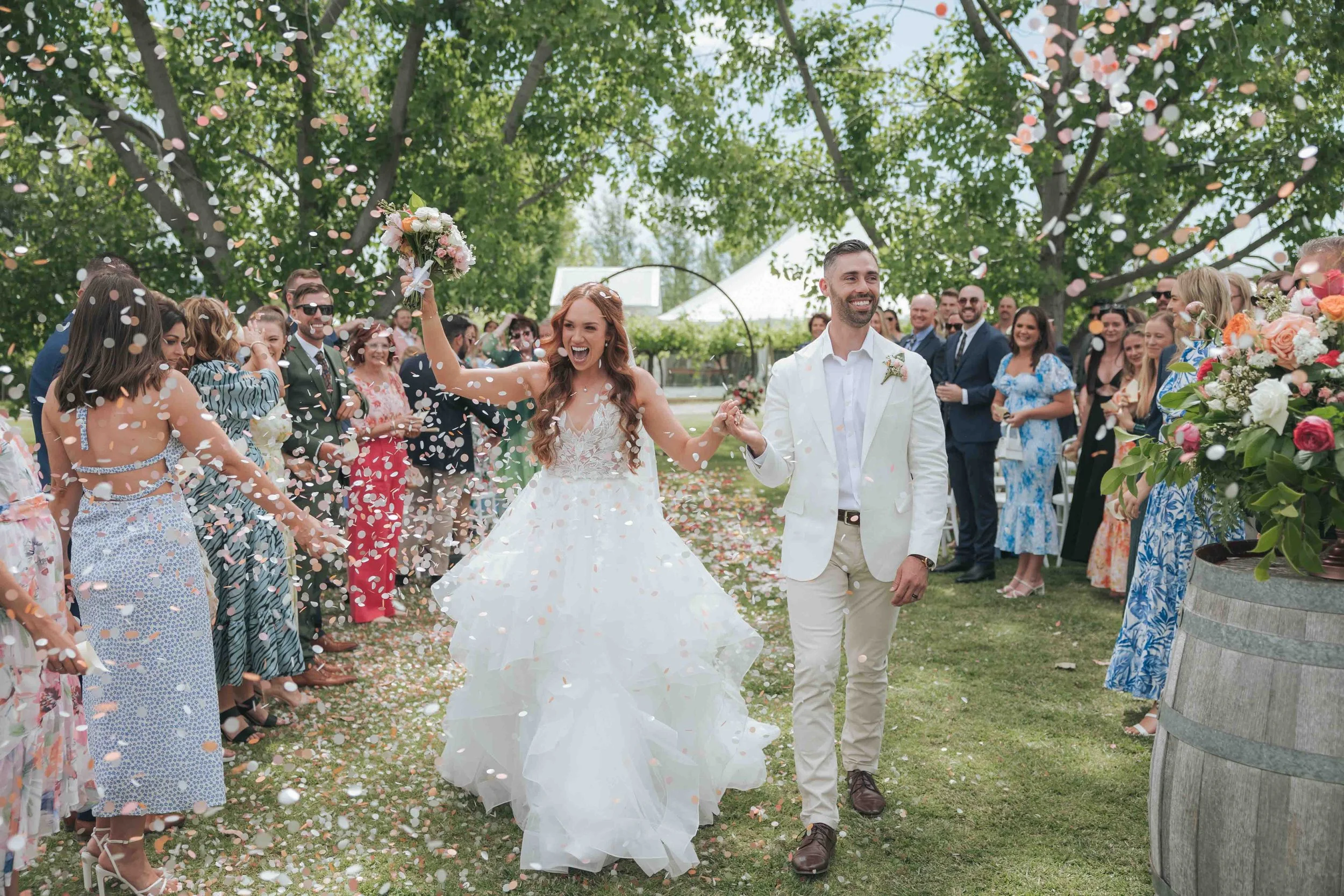 A newlywed couple smiling as they walk through a celebration with guests throwing confetti outdoors under trees.