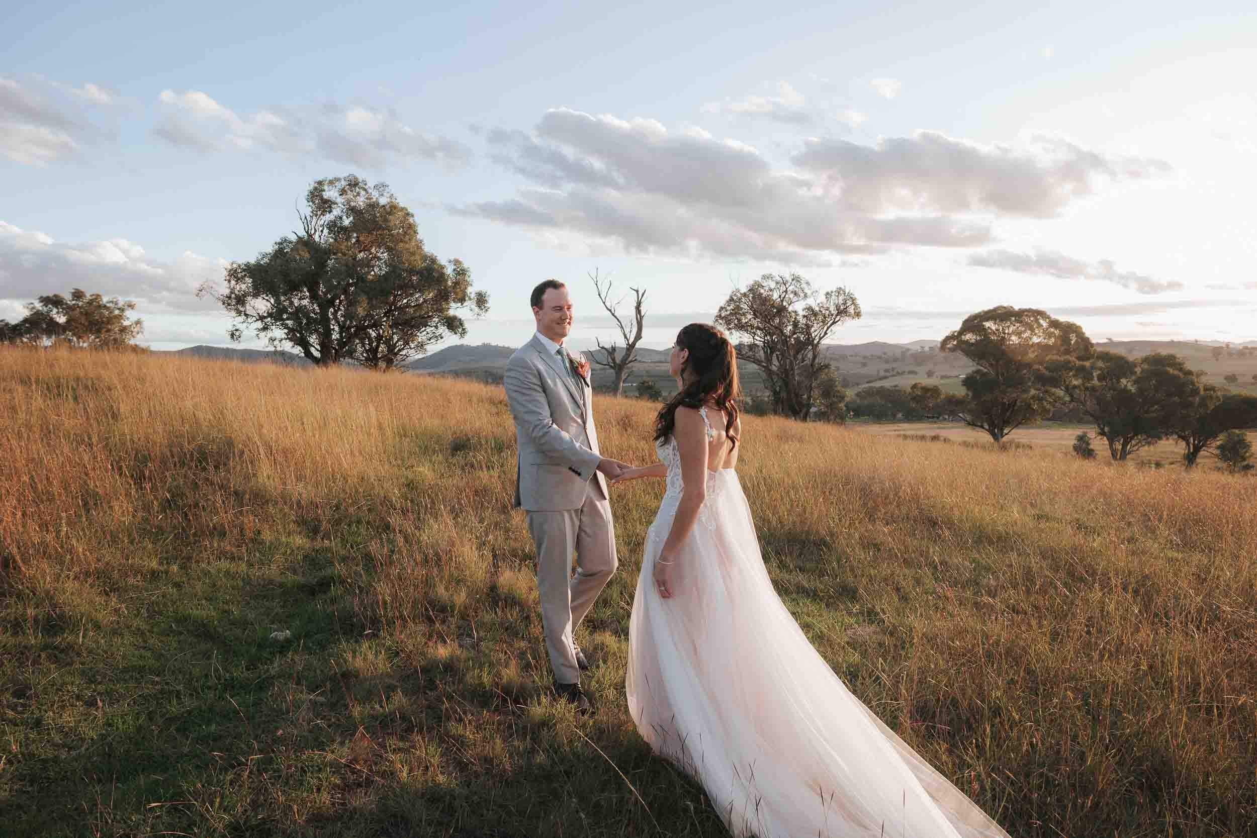 A couple on their wedding day standing in a field of tall grass with trees and distant hills, holding hands and smiling at each other during sunset.