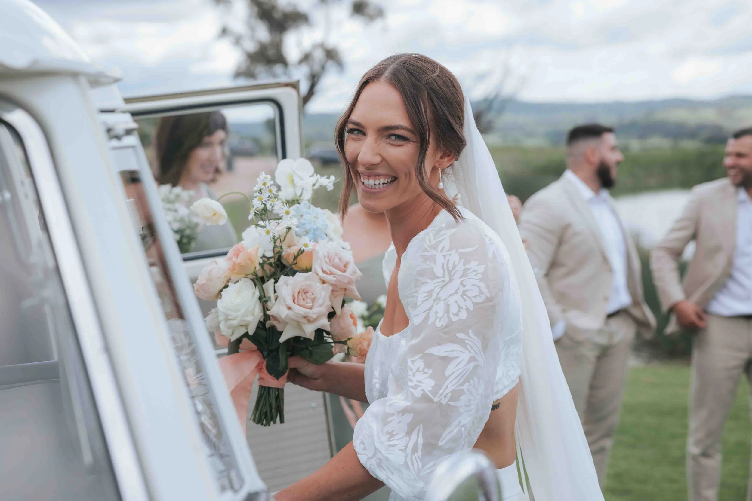 Happy bride in a white dress holding a bouquet of flowers near a vintage auto at an outdoor wedding.