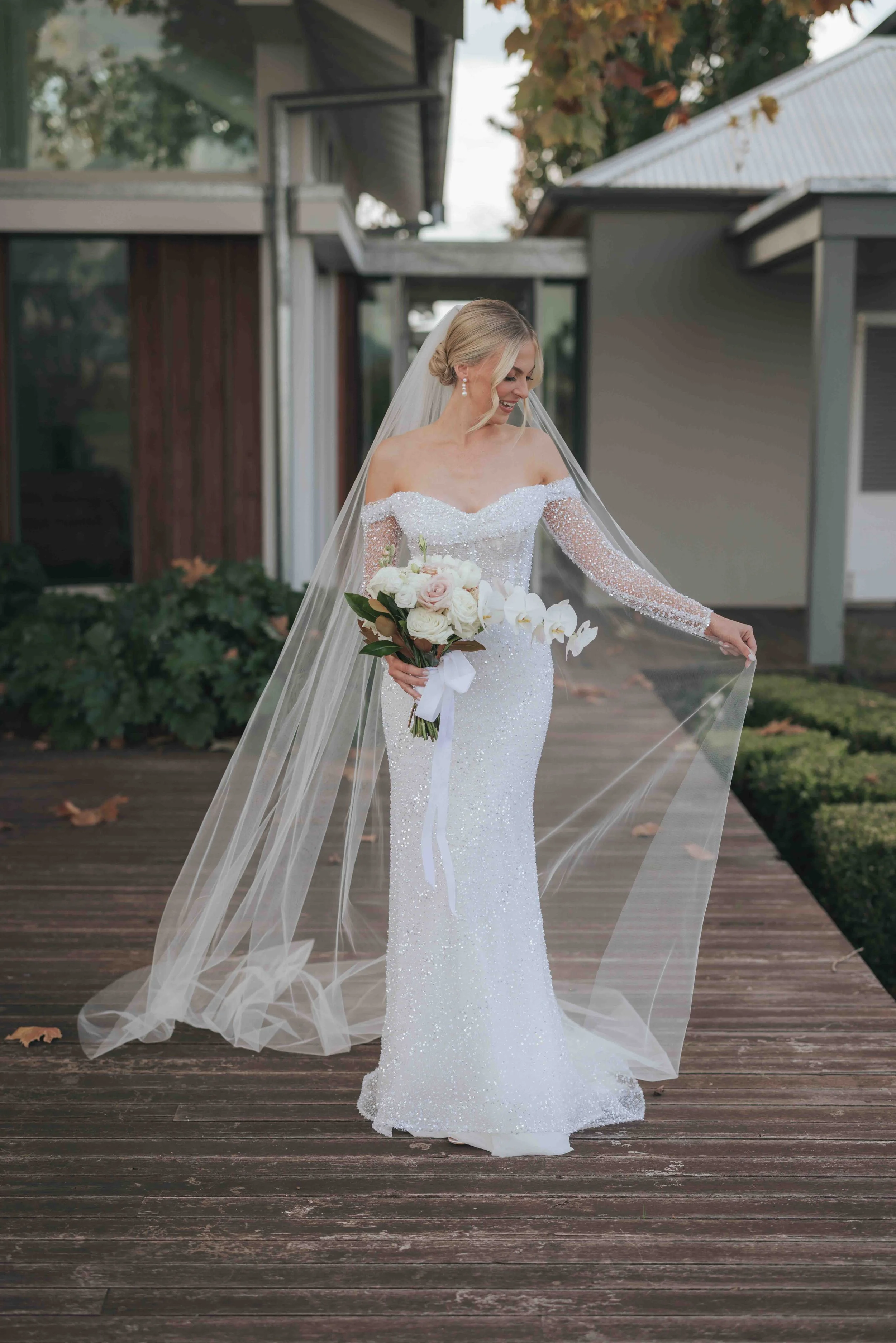 A bride in a white, sparkly wedding gown holding a bouquet of roses and orchids, standing on a wooden deck outdoors, smiling while adjusting her veil.