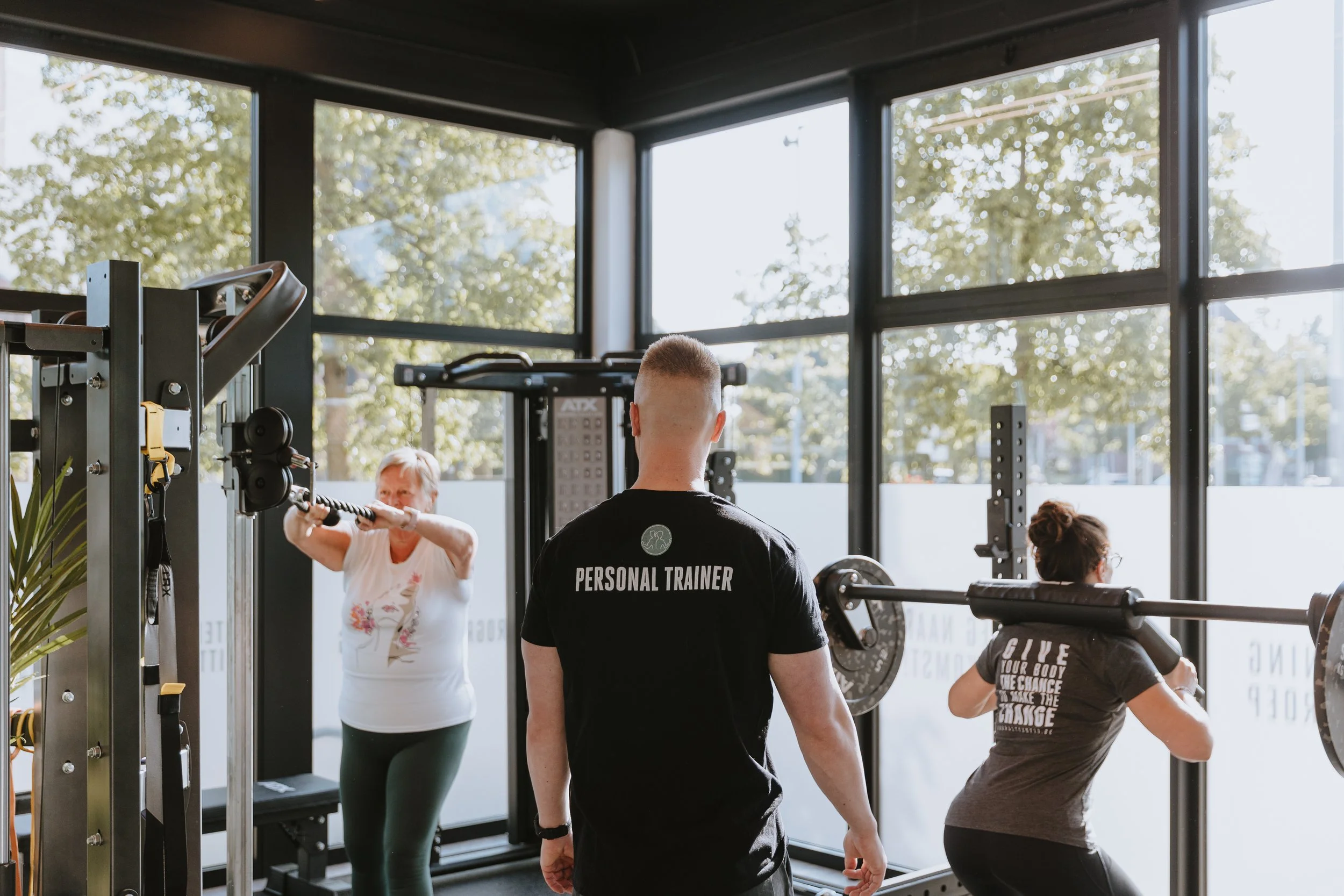 Personal trainer observeert twee vrouwen die krachttraining doen in een sportschool met grote ramen en groene bomen buiten.