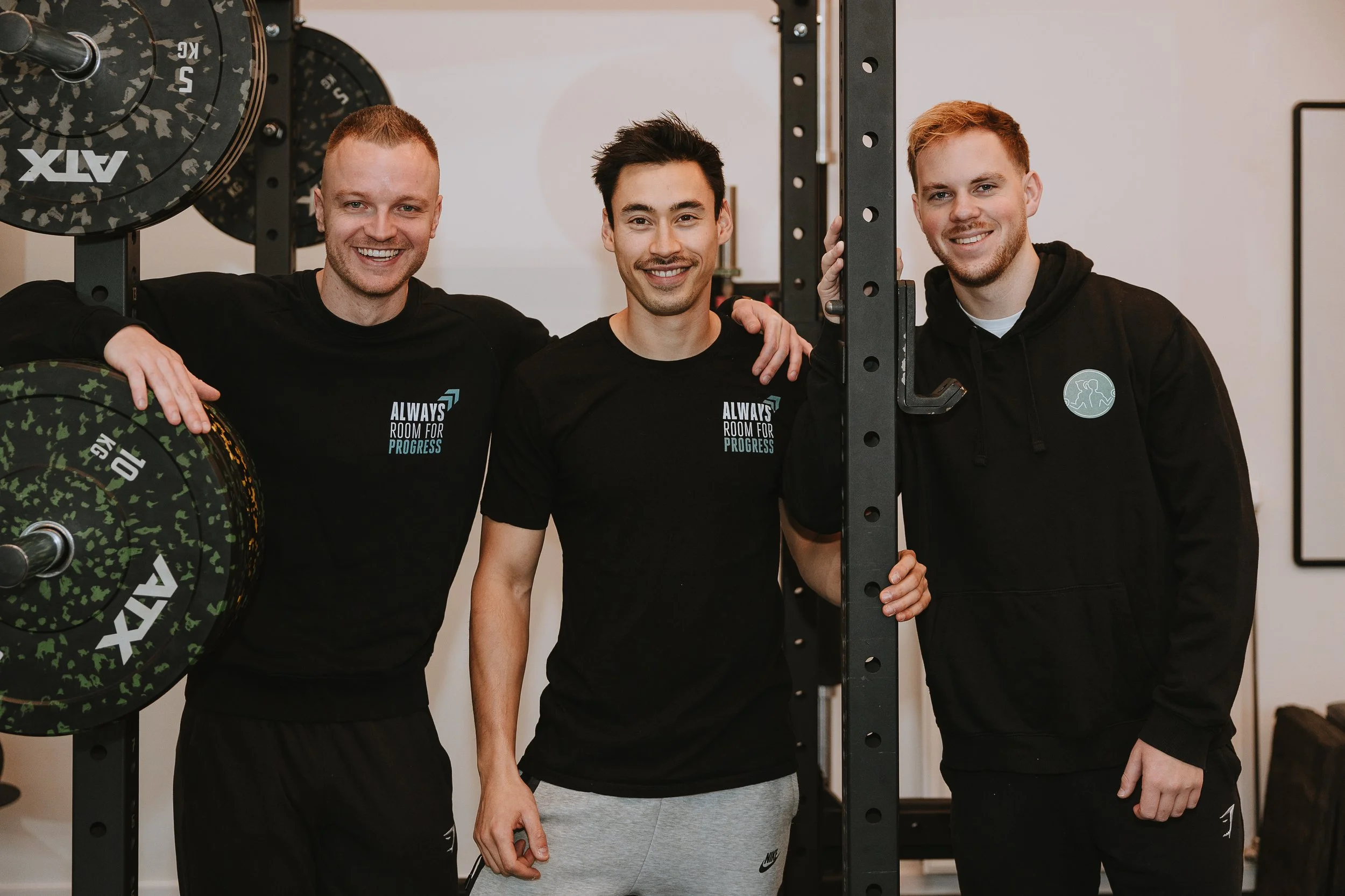 Drie jonge mannen in sportkleding staan samen in een sportschool, glimlachen en poseren voor de camera, met gewichten op de achtergrond.
