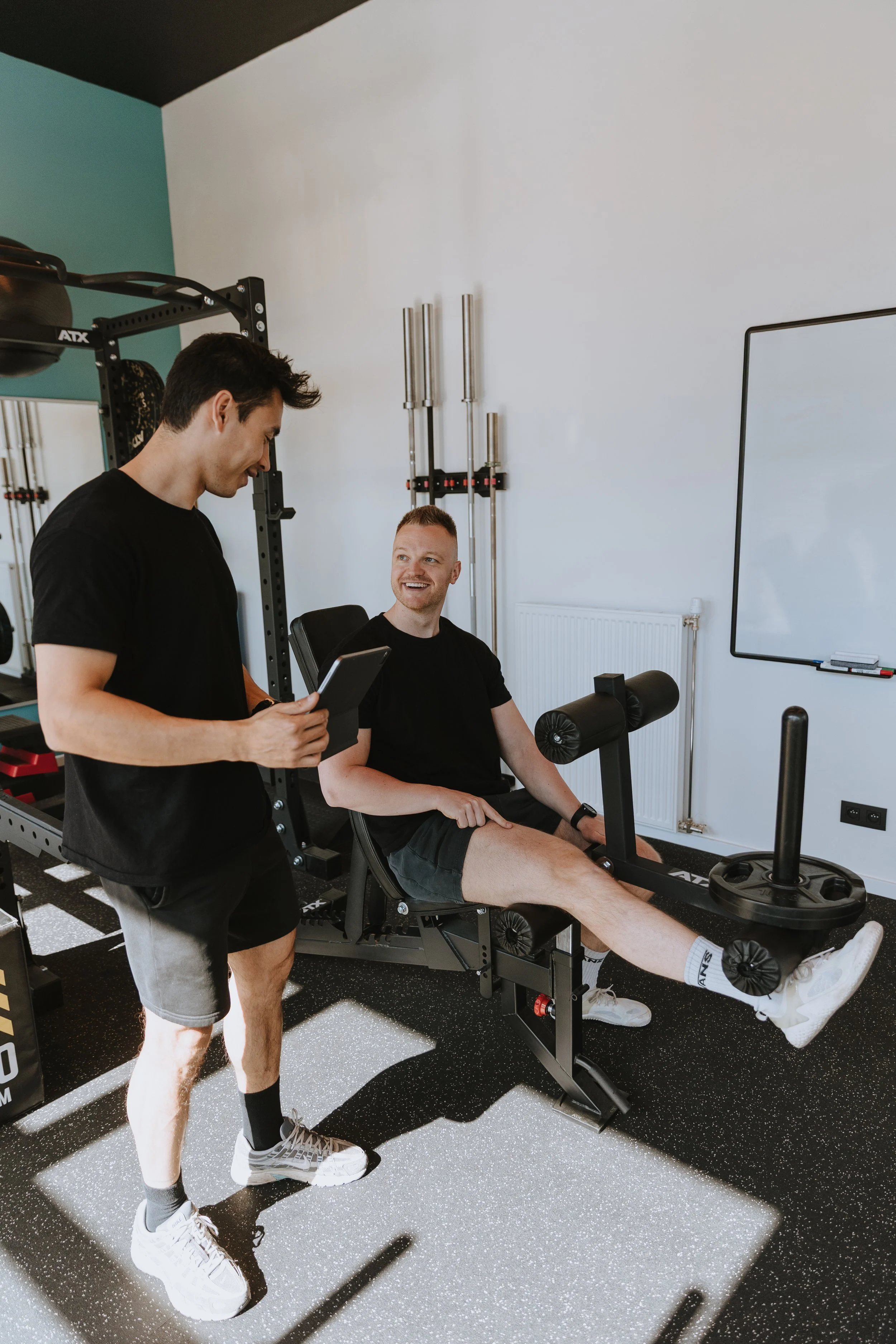 Twee mannen in een fitnessruimte, één zit op een fitnessapparaat en de andere staat ernaast, beide lachen en praten.