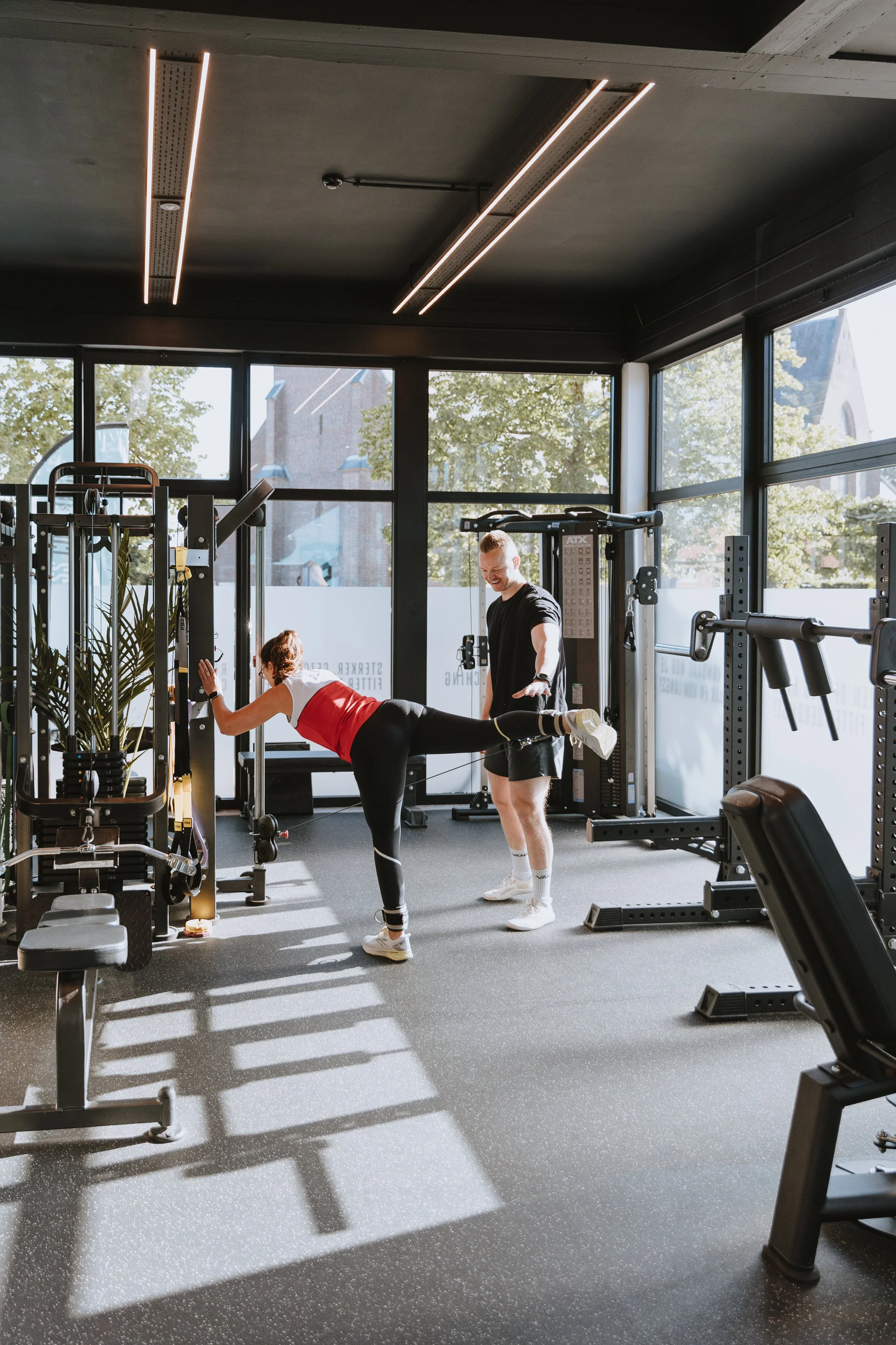 Een persoon die een groepsfitnessles doet onder begeleiding van een instructeur in een nieuwe fitnessruimte met grote ramen en moderne apparatuur.