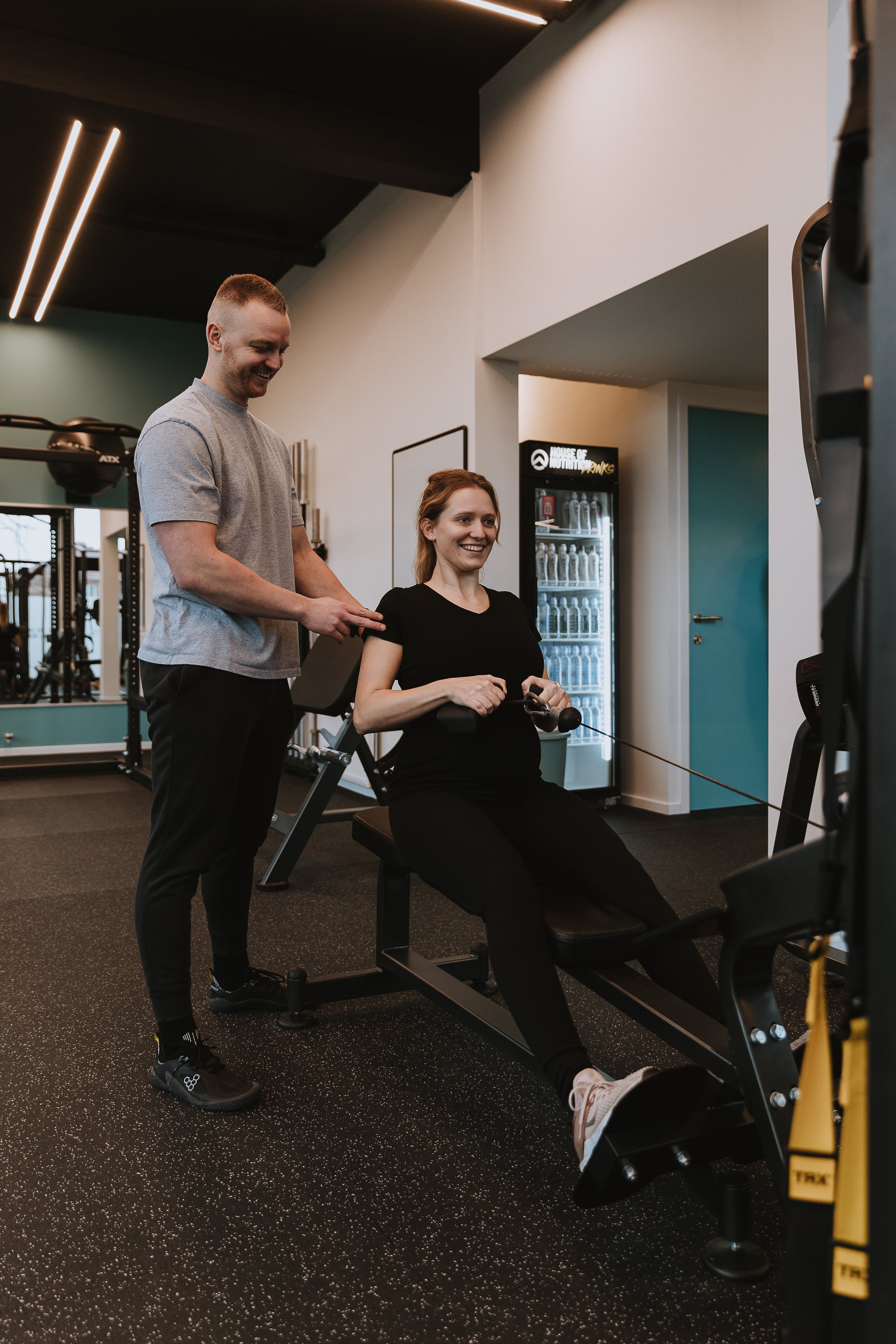 Vrouw doet aan krachttraining op een fitnessapparaat, begeleid door een instructeur in een sportschool.