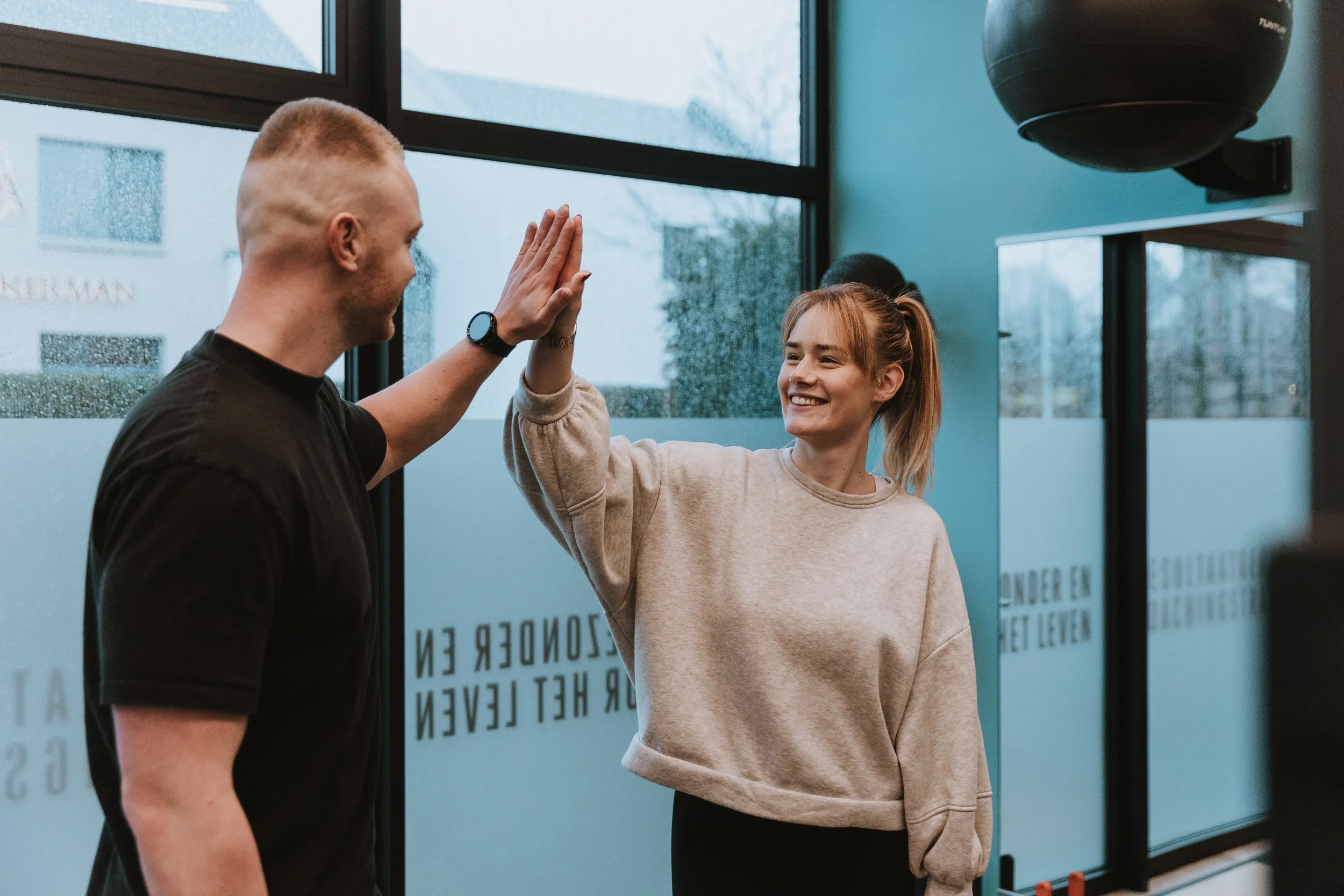 Een man en een vrouw geven elkaar een high-five in een sportschool met grote ramen en ronde trainerringen aan het plafond.