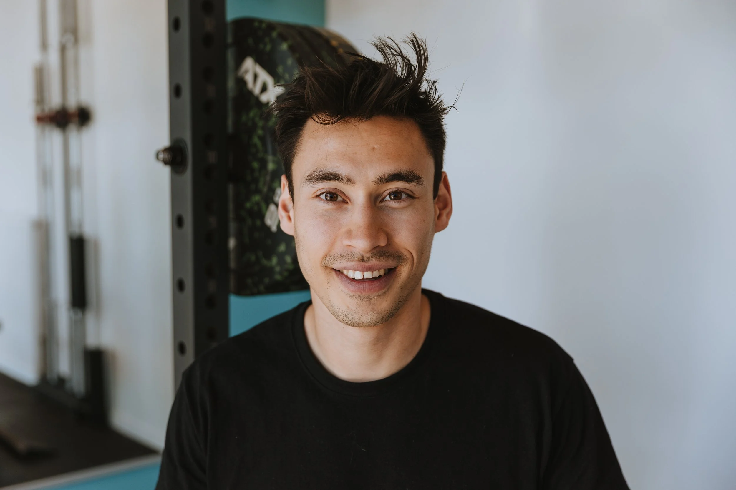 Portret van een lachende jonge man met donker haar, gekleed in een zwarte T-shirt, binnen, met fitnessapparatuur op de achtergrond.