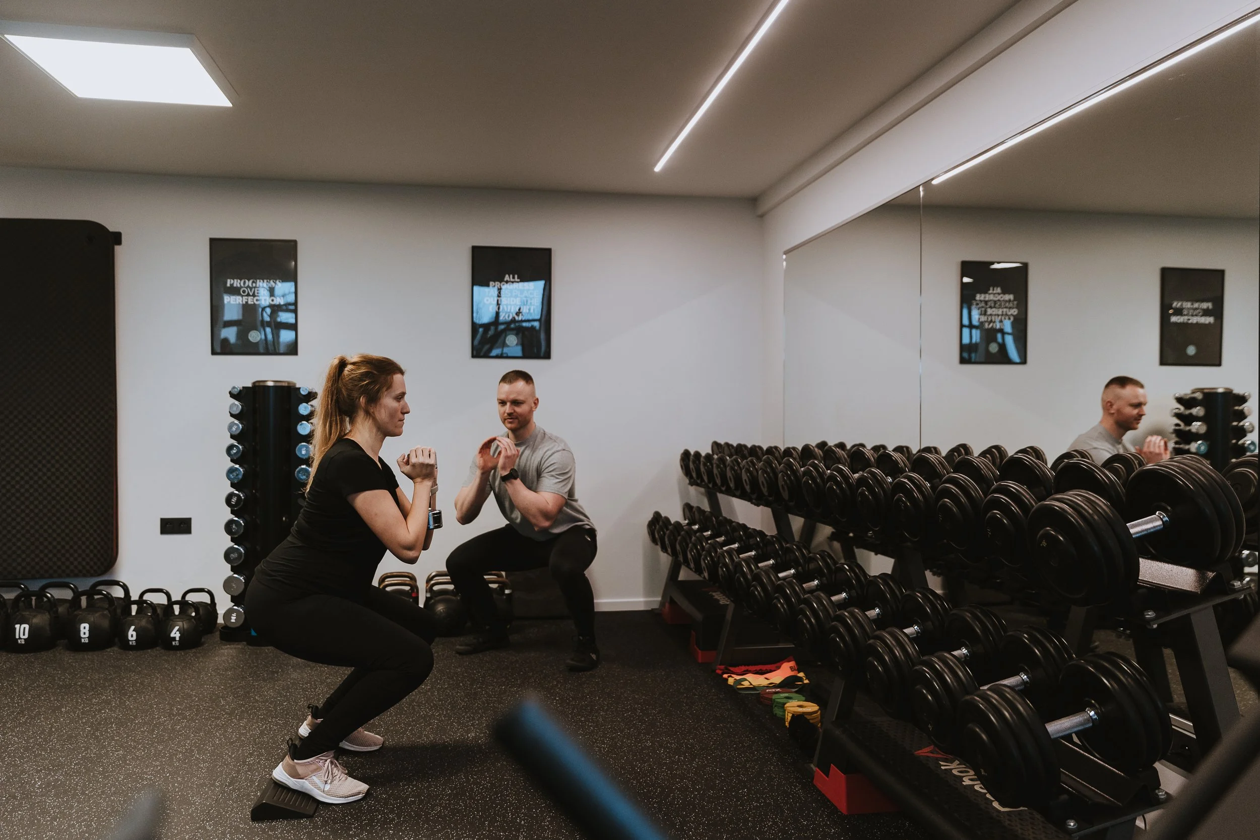 Een personal trainer die in een fitnessruimte oefeningen voor de beenspieren geeft, met gewichten en kettlebells op de achtergrond.