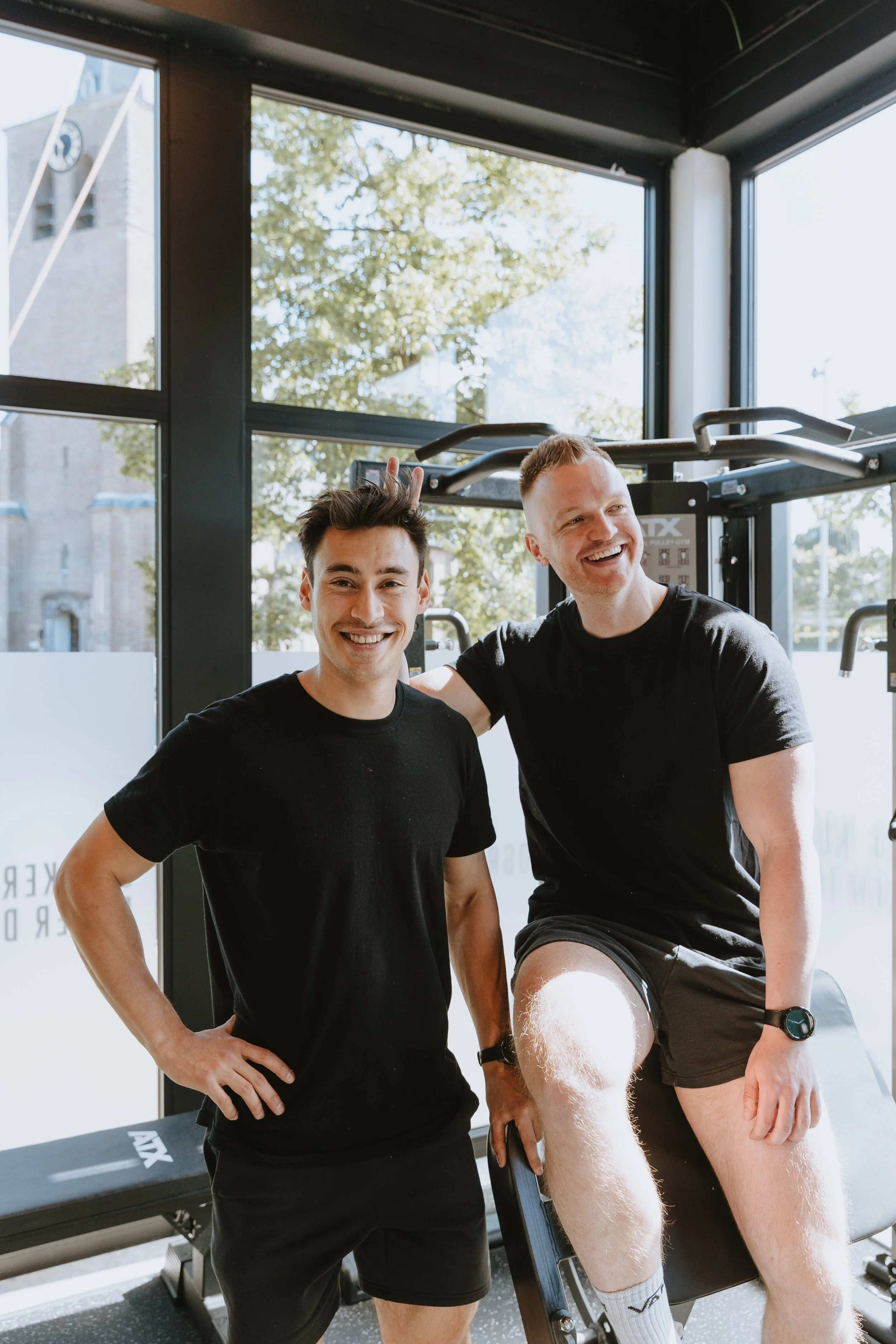 Twee mannen in een sportschool, een glimlacht en lachen, een staat met hand op de heup en de andere op een apparaat, de ander staat naast hem, beide dragen zwarte t-shirts en shorts, in een ruimte met grote ramen en fitnessapparatuur.