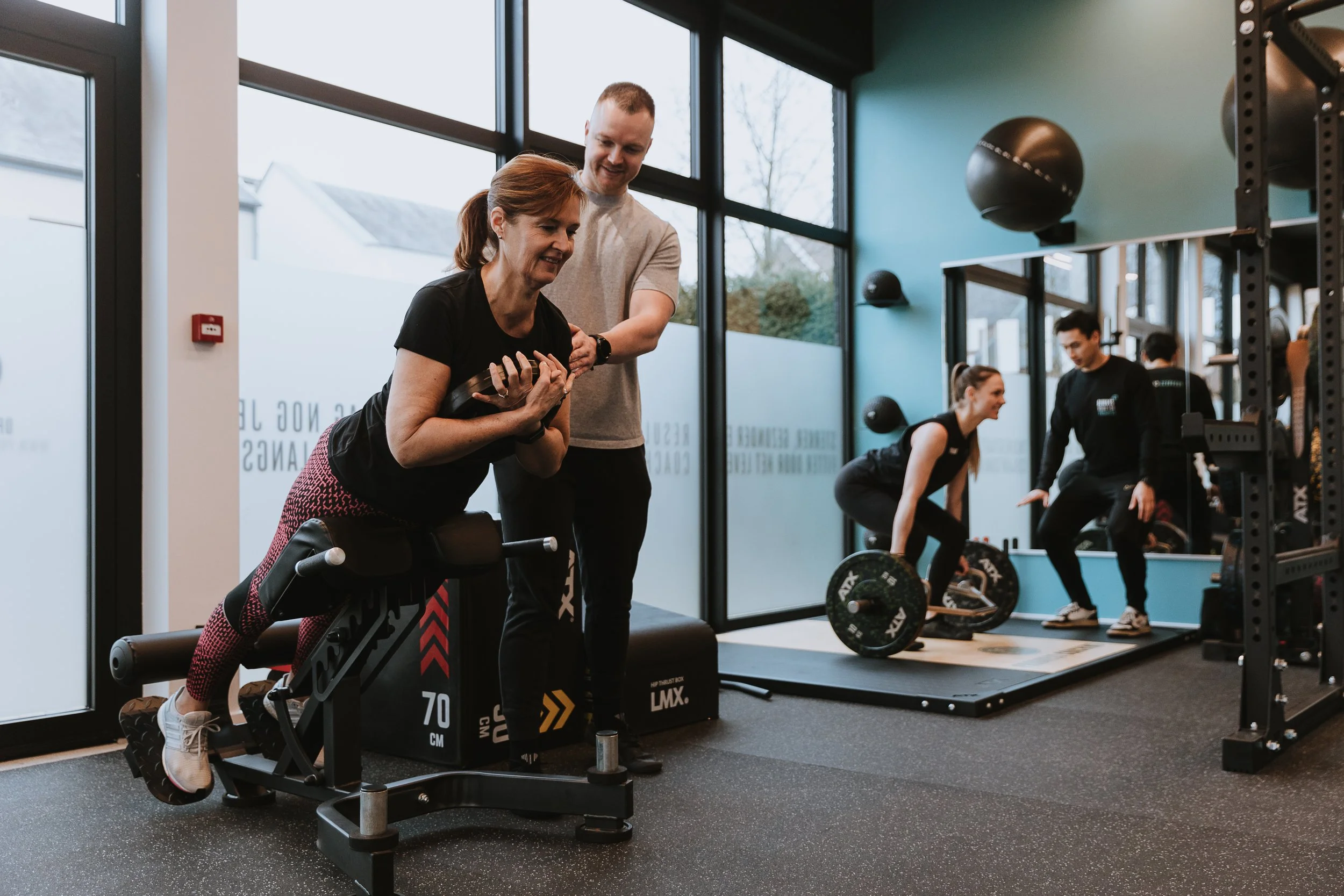 Mensen die krachttraining doen in een sportschool, inclusief een vrouw die op een bankje ligt en een persoon die haar begeleidt, terwijl twee anderen in de achtergrond oefeningen met halters uitvoeren.
