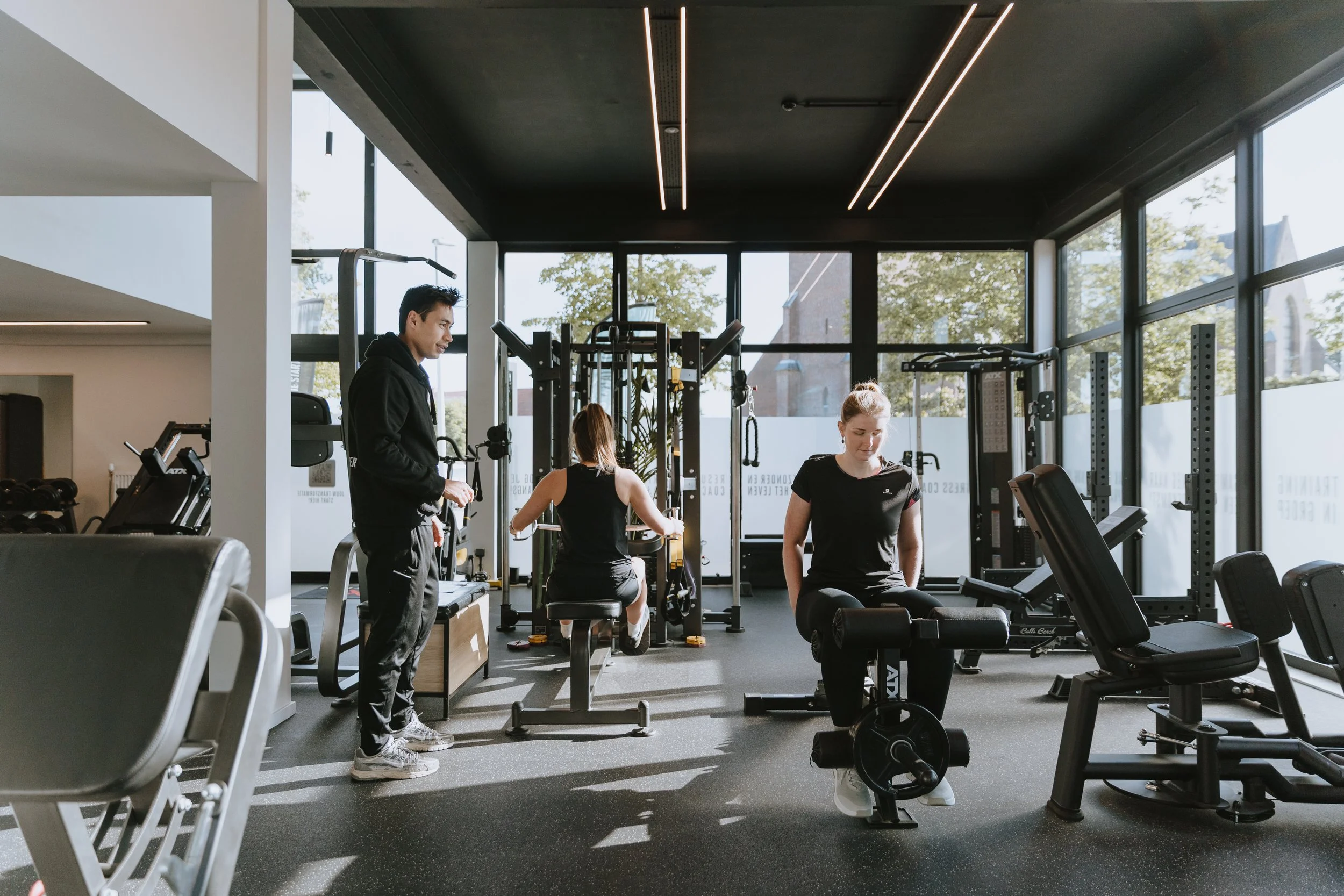 Drie mensen trainen in een moderne sportschool met grote ramen en natuurlijke lichtinval
