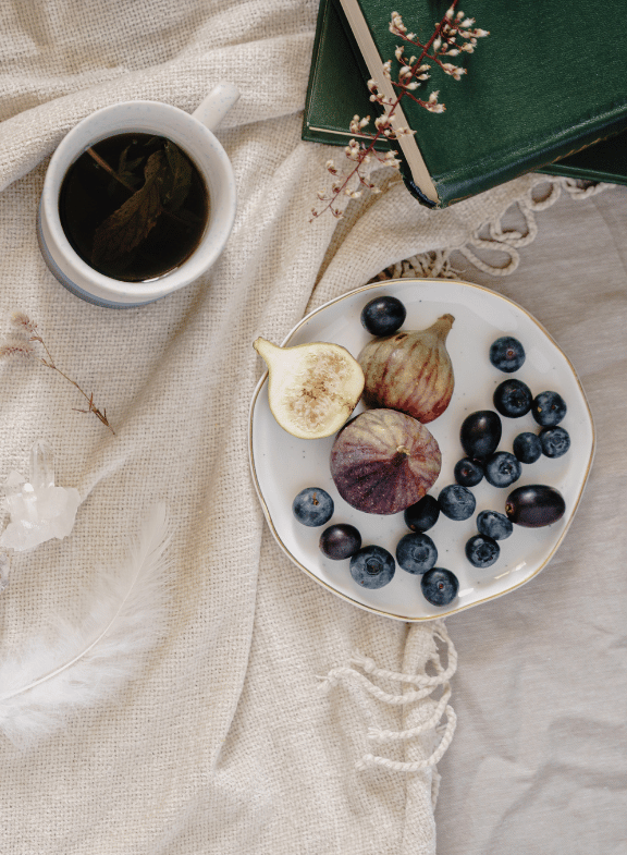 A cup of tea, a plate with figs and blueberries, two green books with a sprig of small white flowers, and a feather on a cream-colored fabric surface.