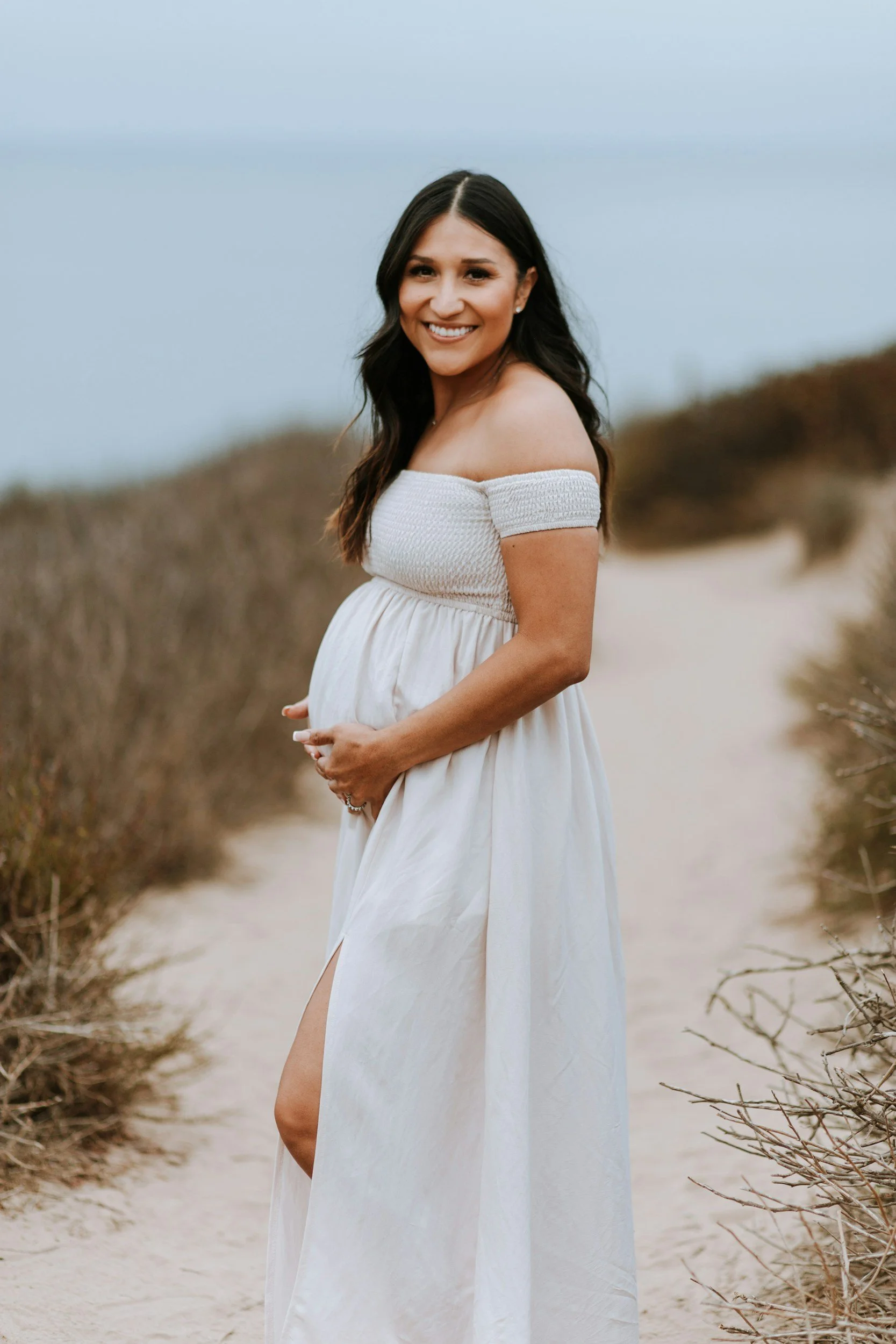 pregnant woman wearing white flowing dress holding her belly on white sand path