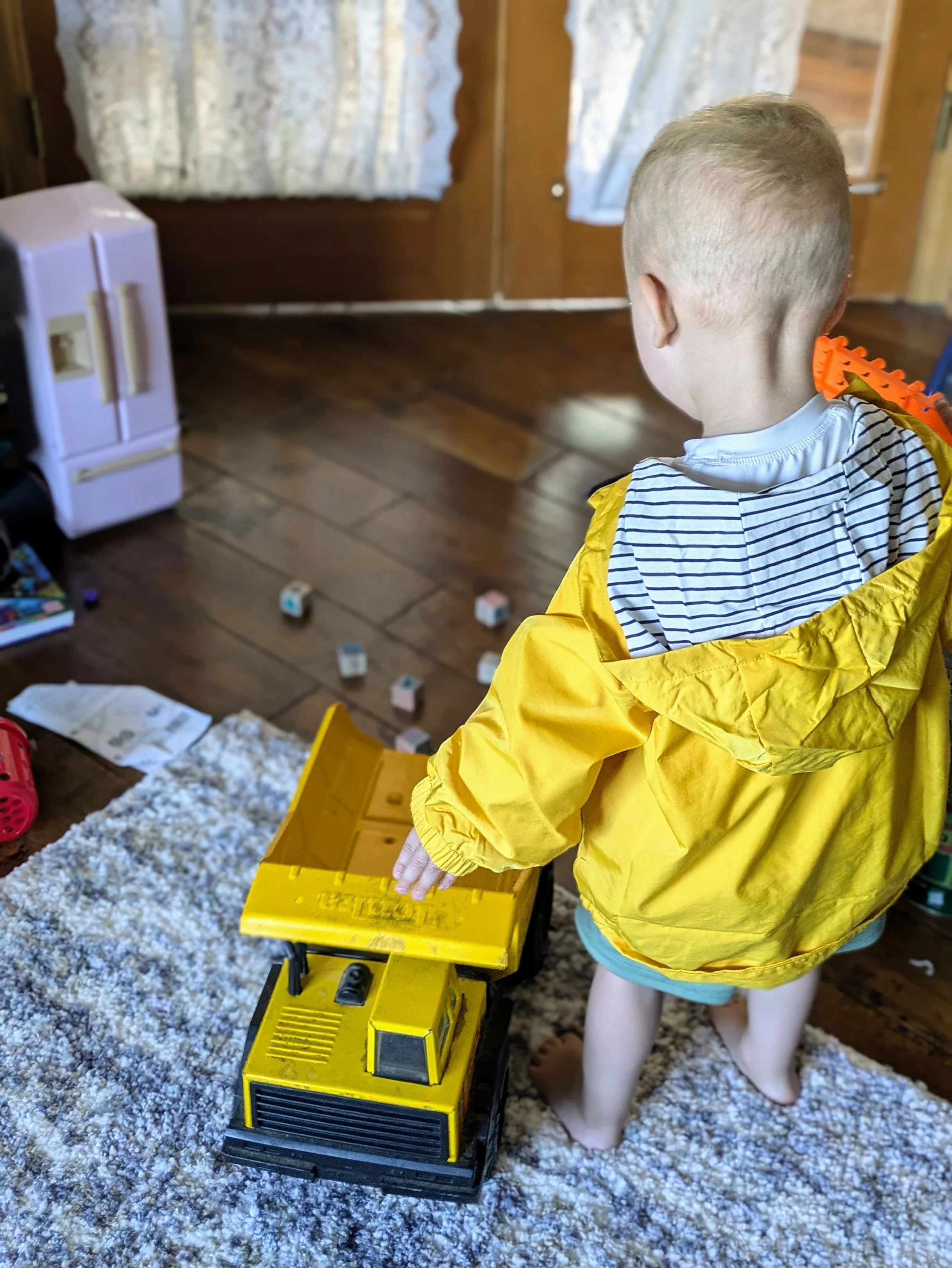 Toddler playing in yellow rain jacket on a rug with an old tonka dump truck.