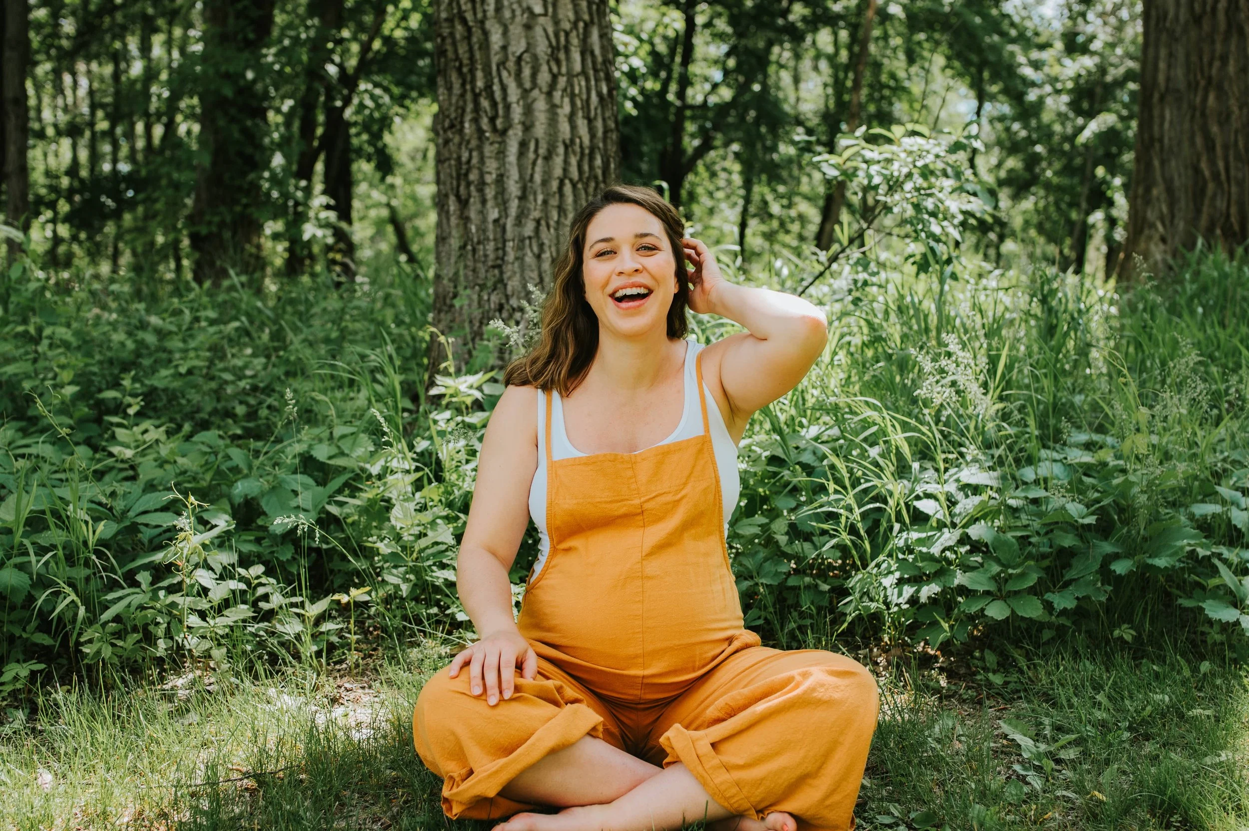 laughing pregnant woman (doula Samantha Ashley) in a golden jumper sitting in green grass with trees and nature behind her.