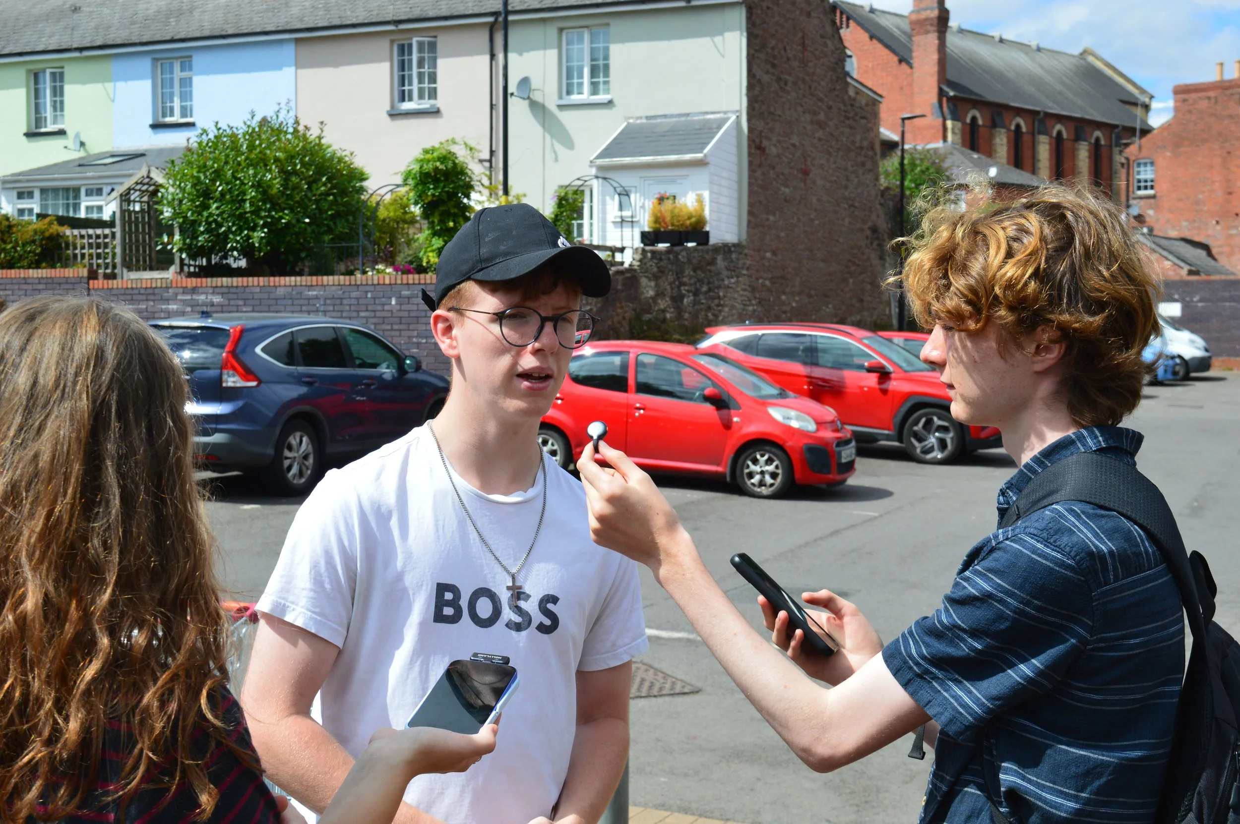 A teenager in a white tshirt and baseball cap is being interviewed on the street by two other young people. The interviewee is speaking into a small microphone held by the interviewer who wears a blue stripey tshirt.