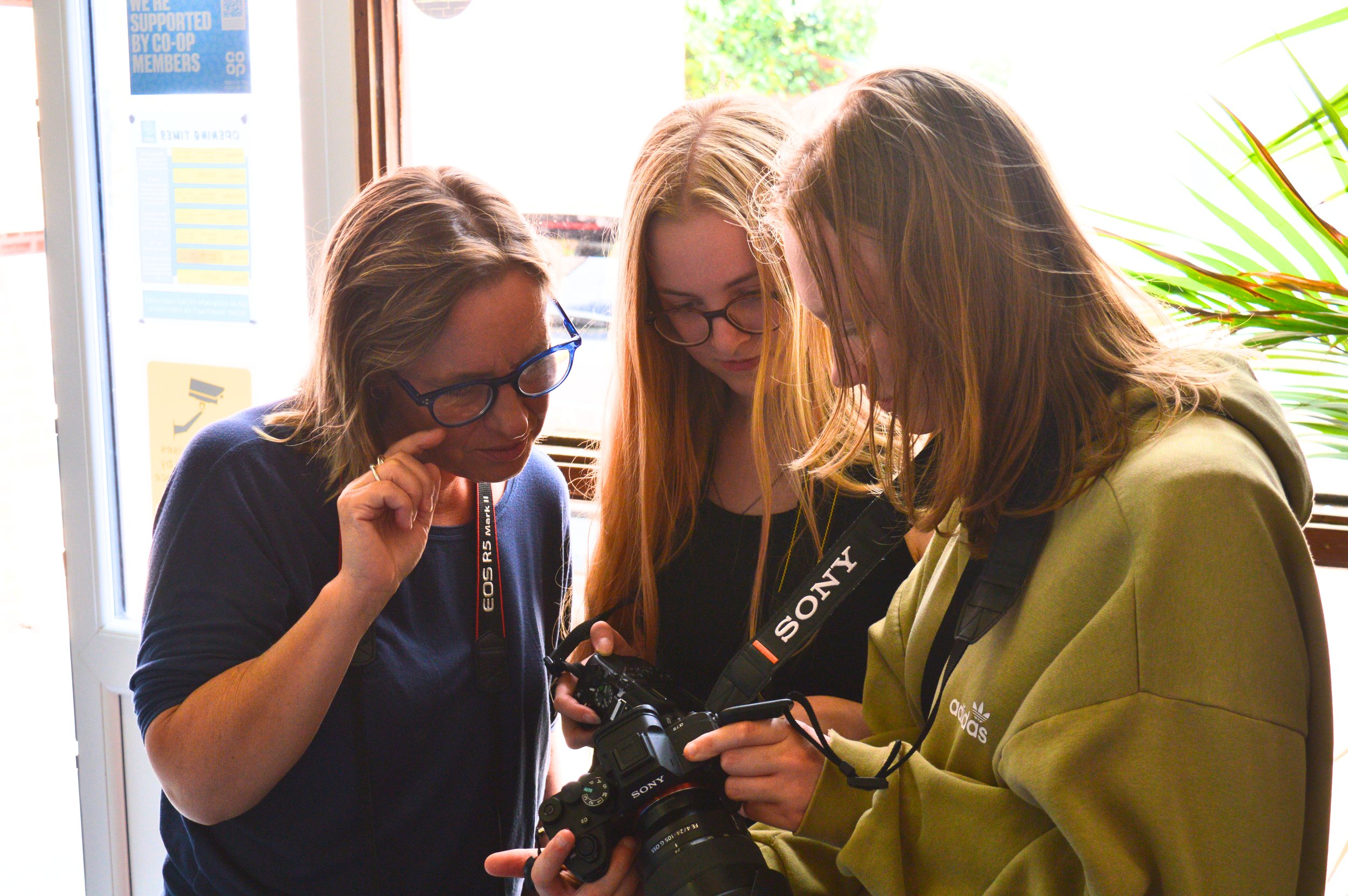 Two young women, in their teens, stand next to each other and both look at the viewing screen of a digital camera. They are with an older woman who is their photograpy tutor.