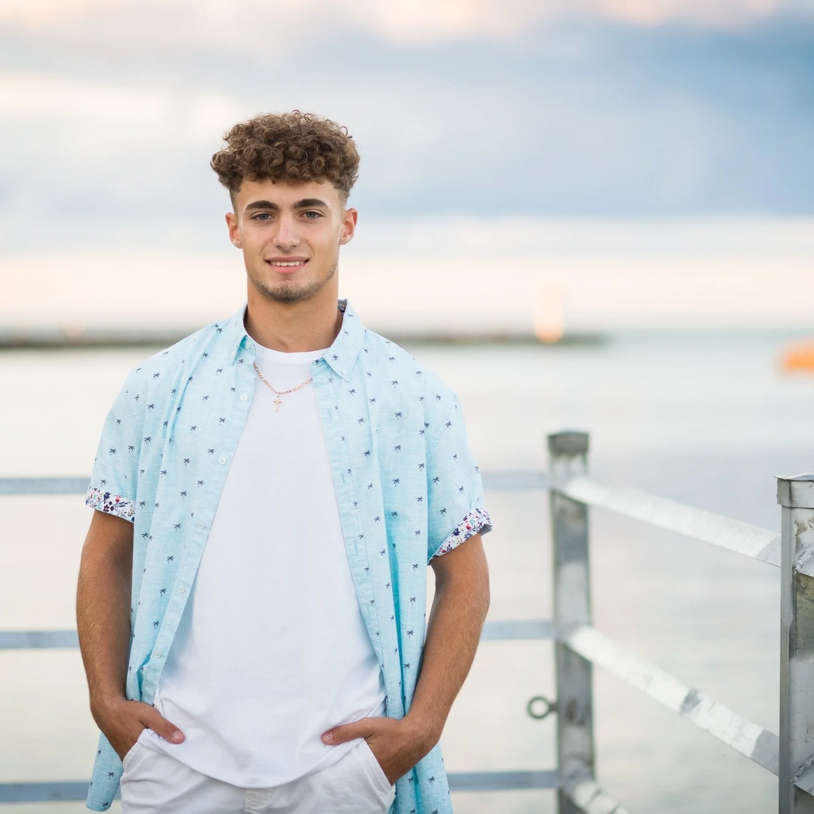 A young man with curly brown hair and light skin standing beside a wooden railing on a waterfront pier during sunset or early evening, wearing a white t-shirt and a light blue, short-sleeved shirt with small palm tree patterns, looking at the camera.