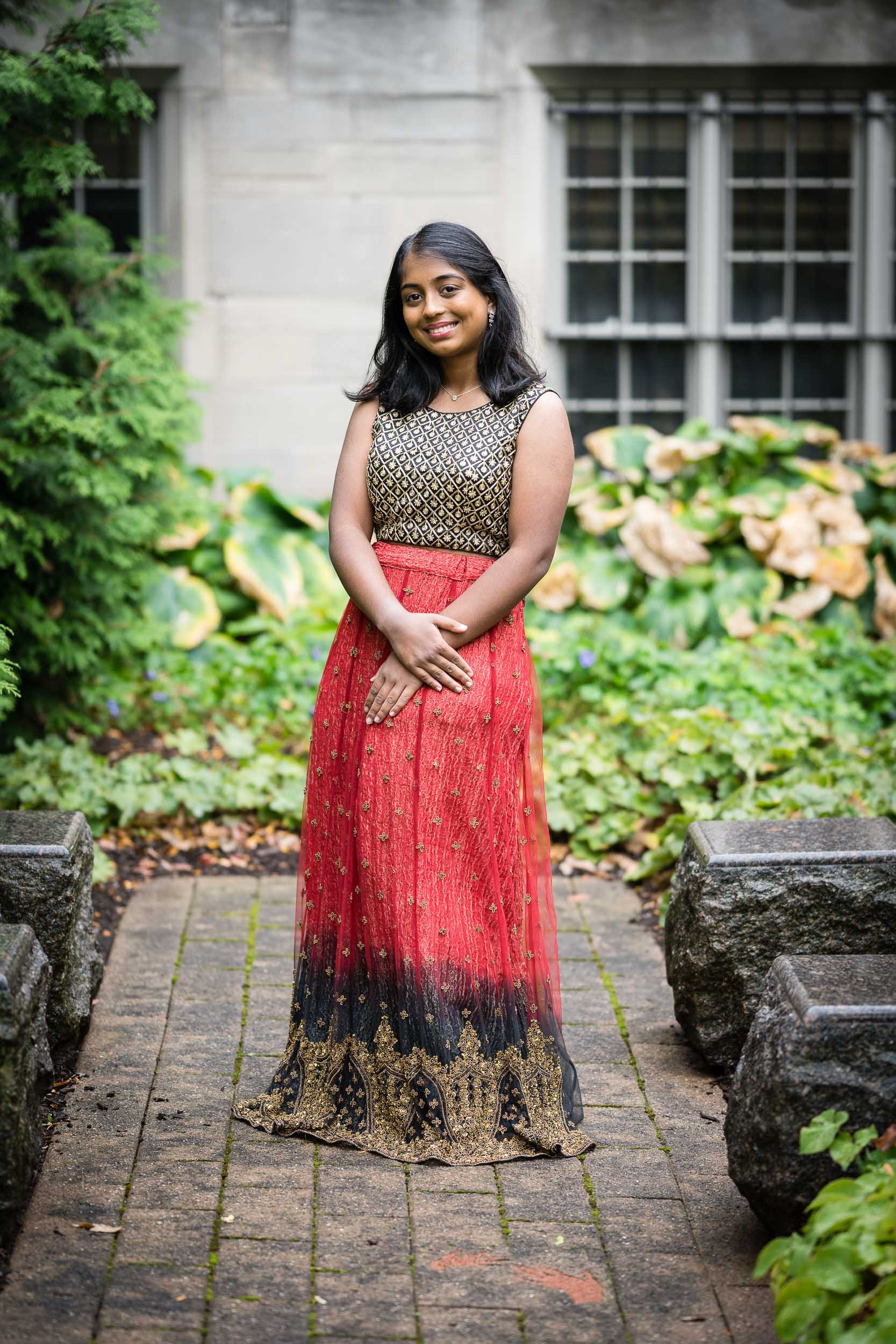 A young woman stands on a stone pathway in front of a gray stone building with large windows, wearing a traditional Indian outfit with a gold and black patterned top and a long red skirt. She is smiling and has shoulder-length black hair.