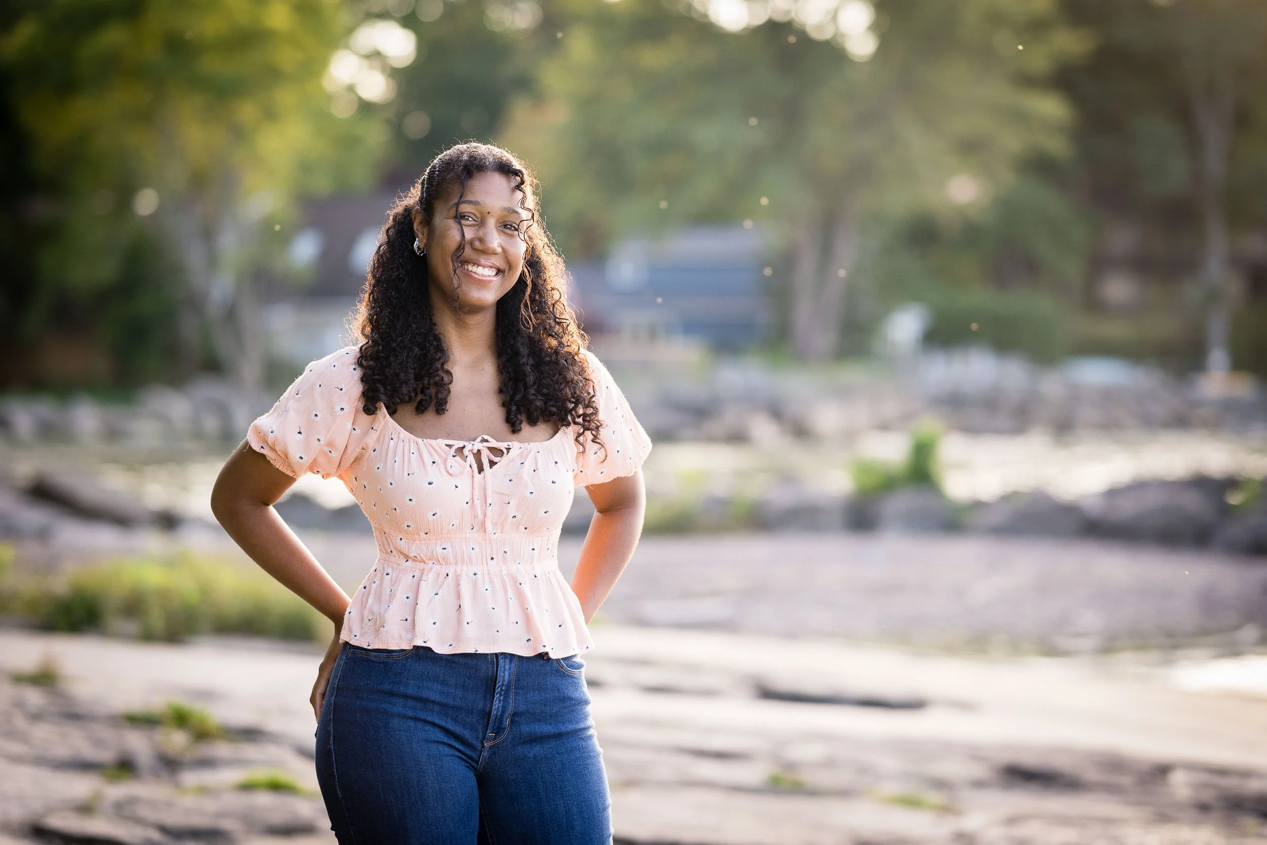 A young woman with curly hair smiling outdoors, wearing a light pink polka dot blouse and blue jeans, with a blurred background of trees and water.