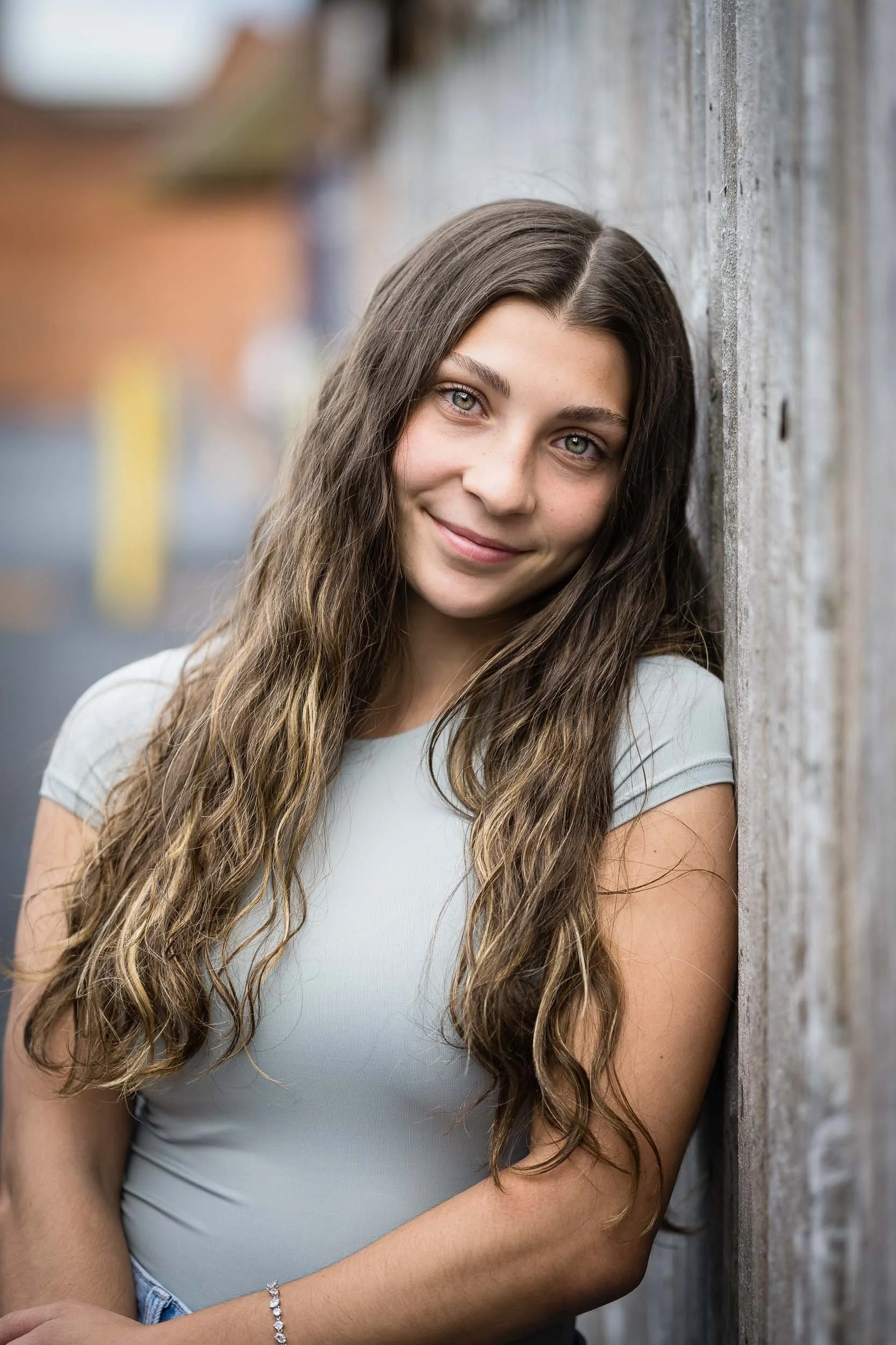 A young woman with long, wavy brown hair and light green eyes leaning against a gray wooden wall, smiling at the camera, wearing a light gray top and a silver bracelet on her wrist.