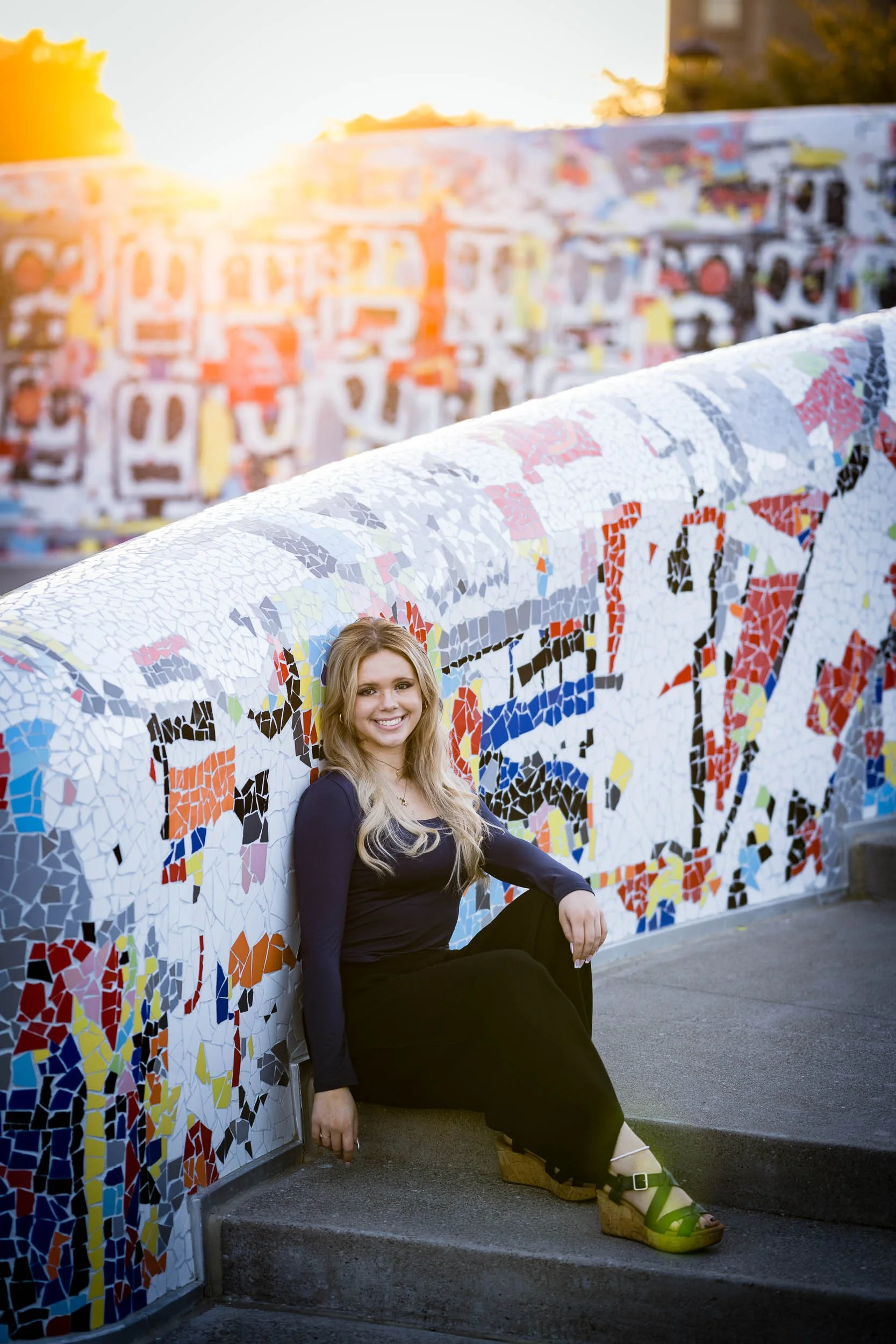A woman with long blonde hair, wearing a black dress and green wedge sandals, sitting on a concrete step next to a colorful mosaic wall during sunset.