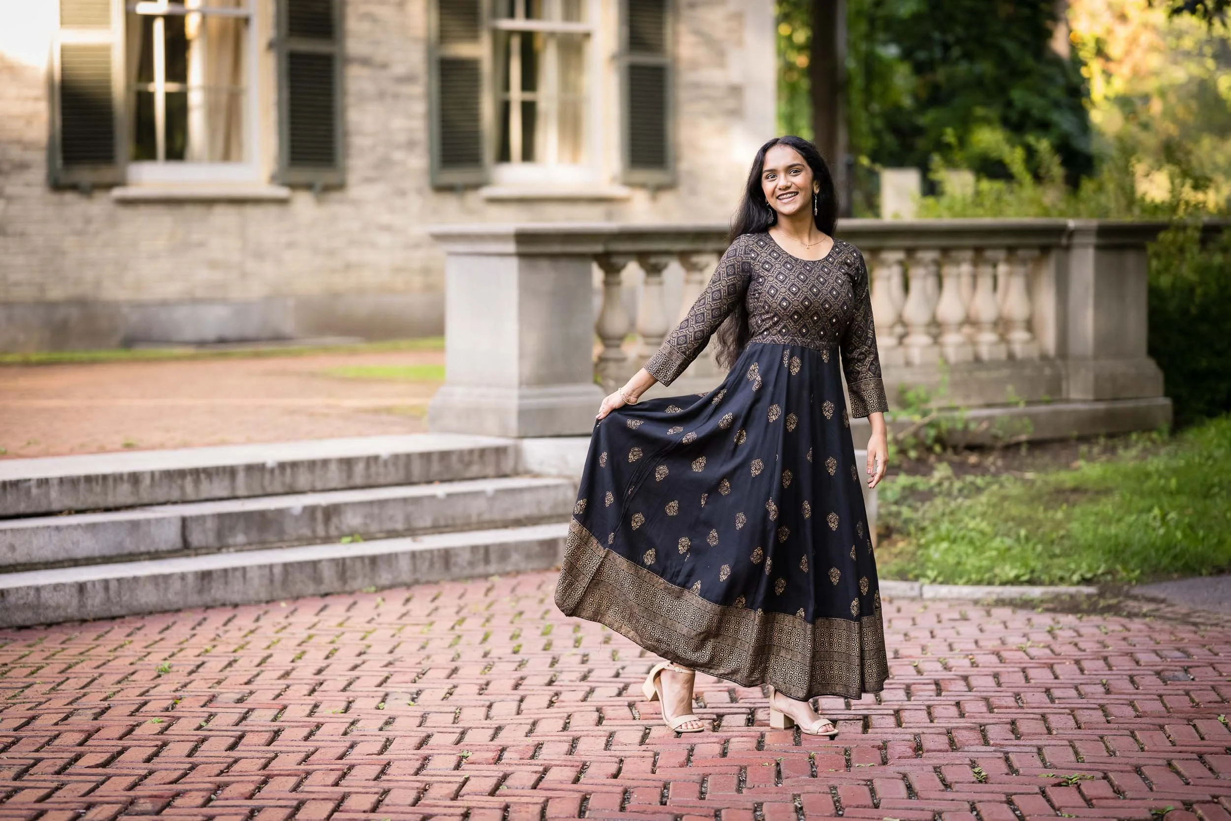 A woman in a traditional dark-colored dress with gold patterns, smiling, standing outdoors on brick steps in front of a stone building with open window shutters and lush green trees in the background.