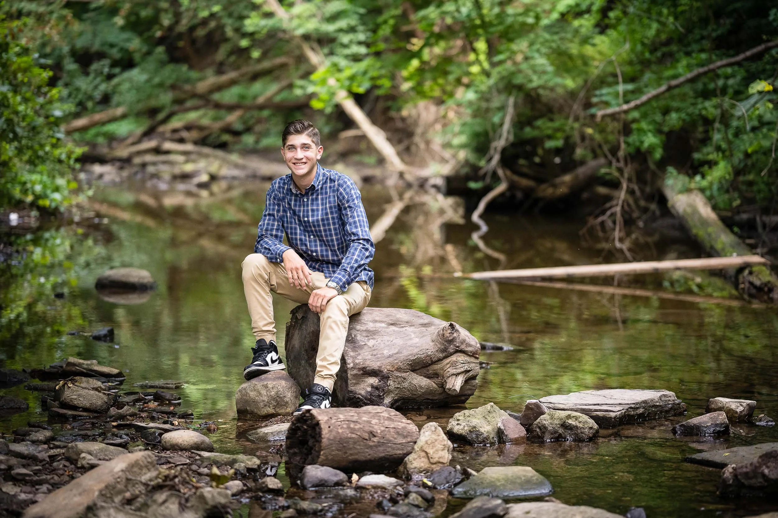 A young man sitting on a large rock in a creek surrounded by dense green trees and foliage.
