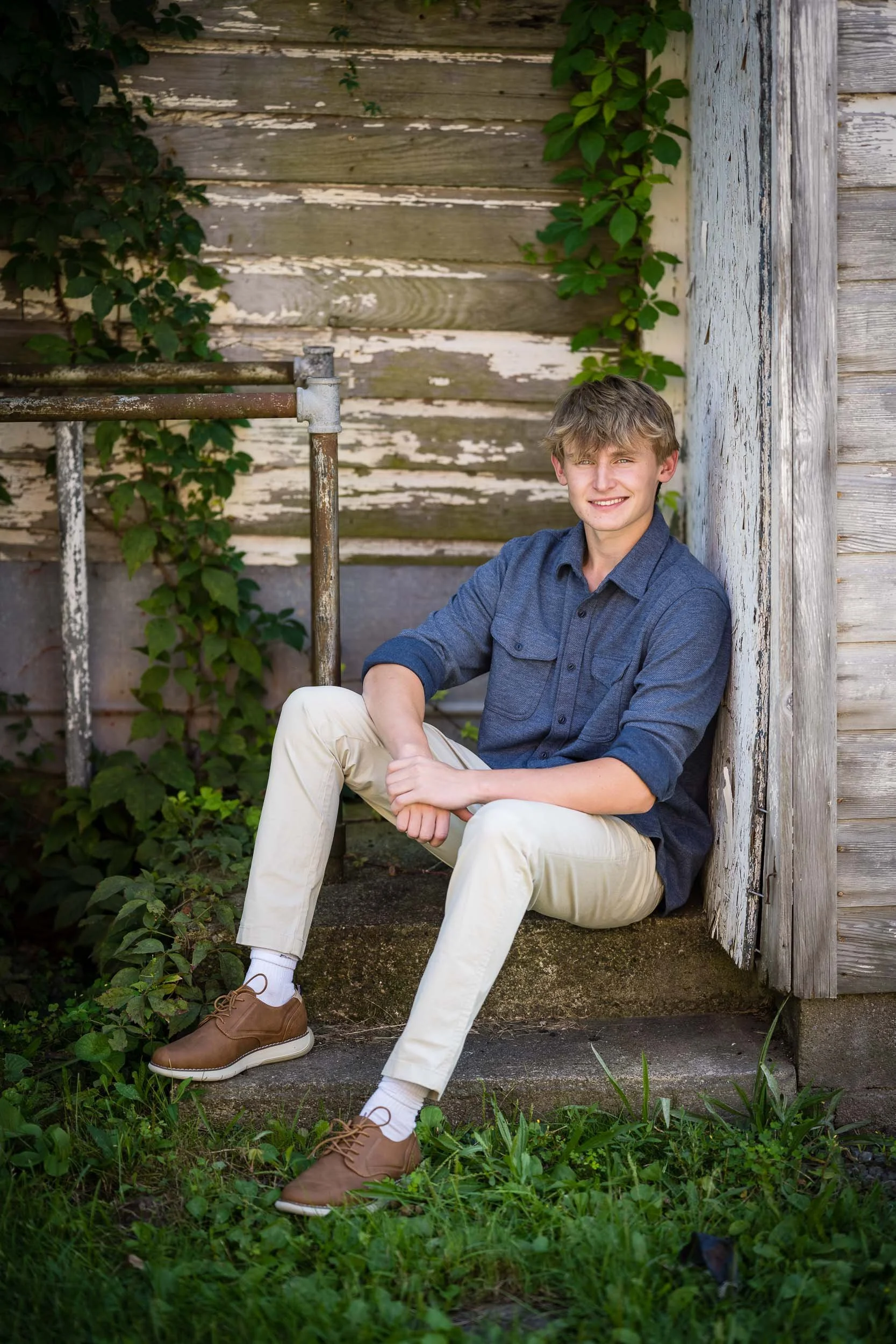 A young man in a blue shirt and cream pants sitting on stone steps, smiling, with a weathered wooden wall and green foliage in the background.