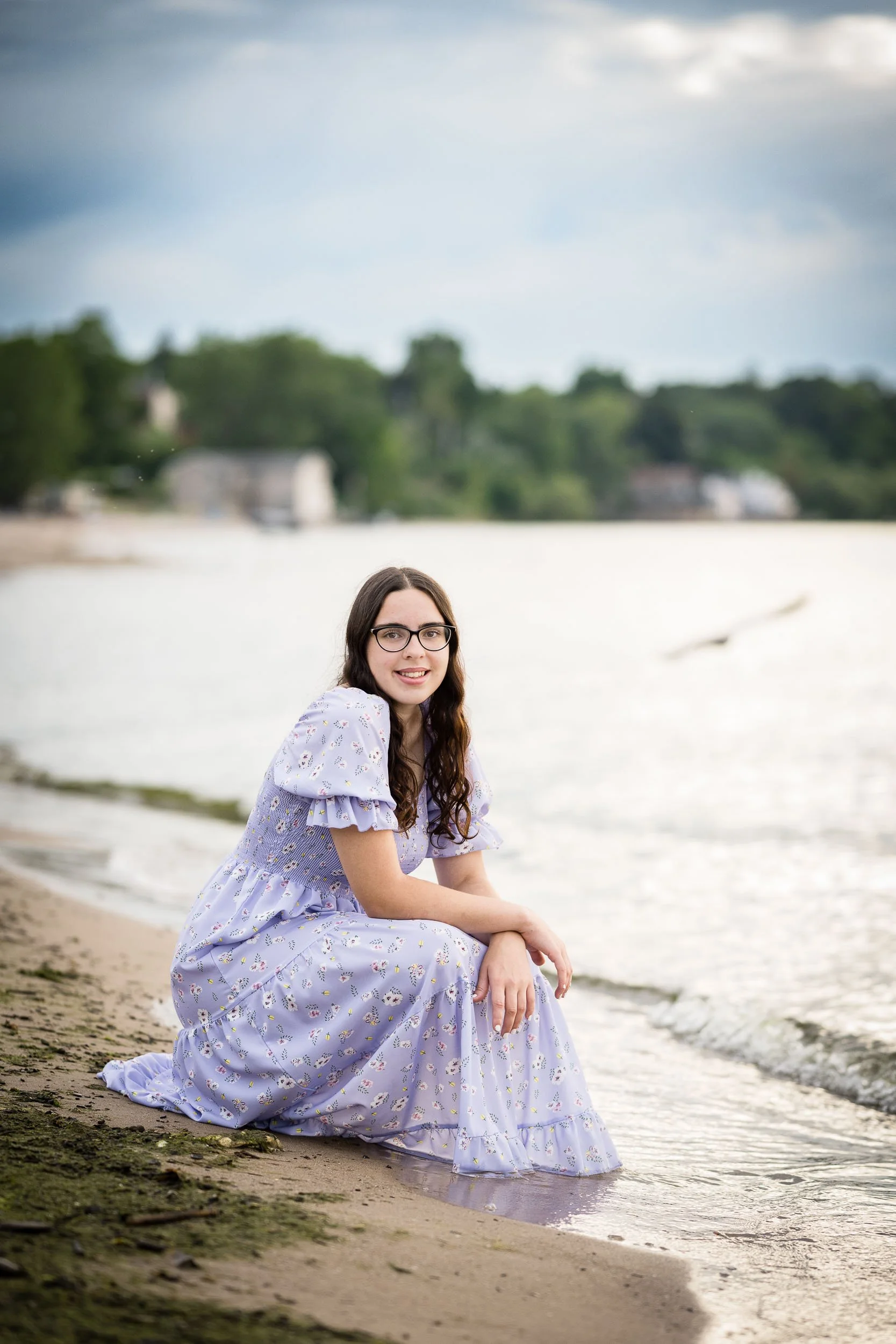 A young woman with dark hair and glasses sitting on the edge of a lake or river, wearing a light purple dress with floral patterns, with trees and cloudy sky in the background.