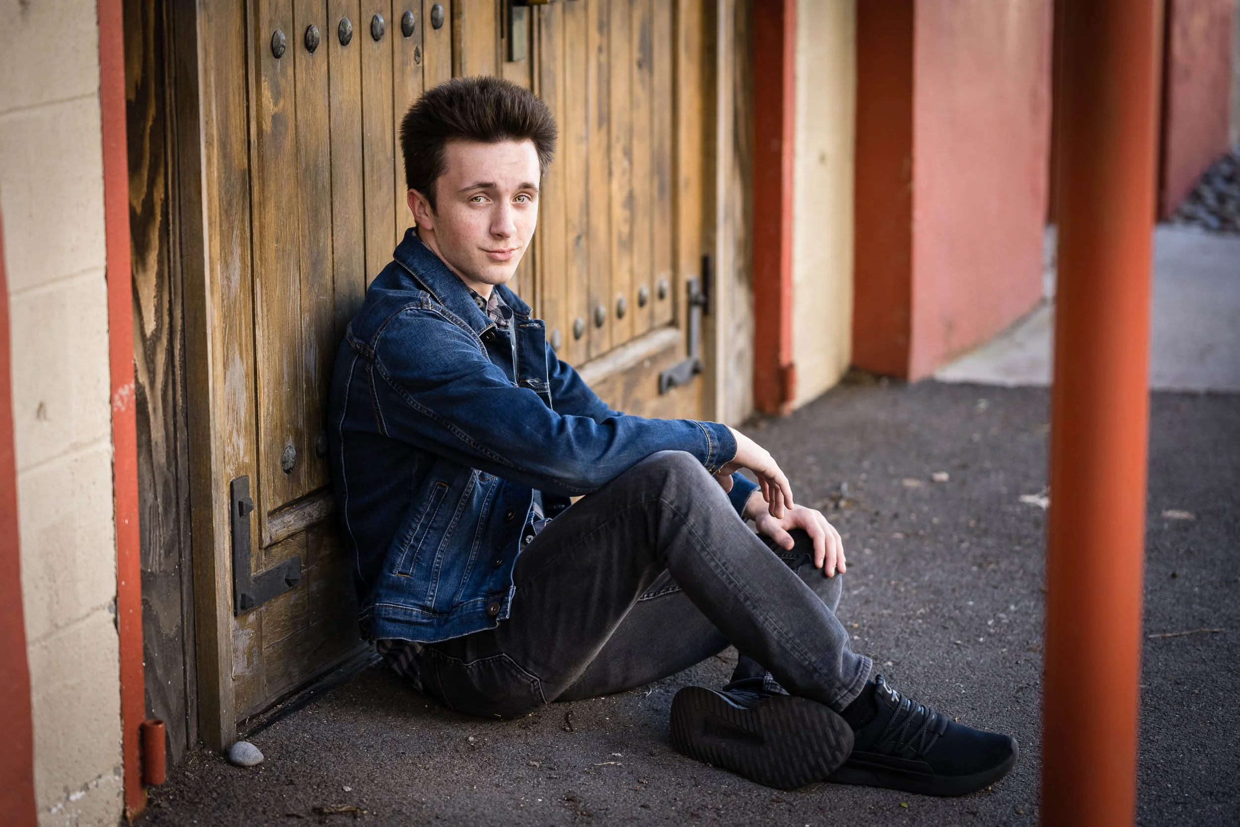 A young man with dark hair, wearing a denim jacket and jeans, sitting on the sidewalk against a wooden door, looking at the camera with a slight smile.