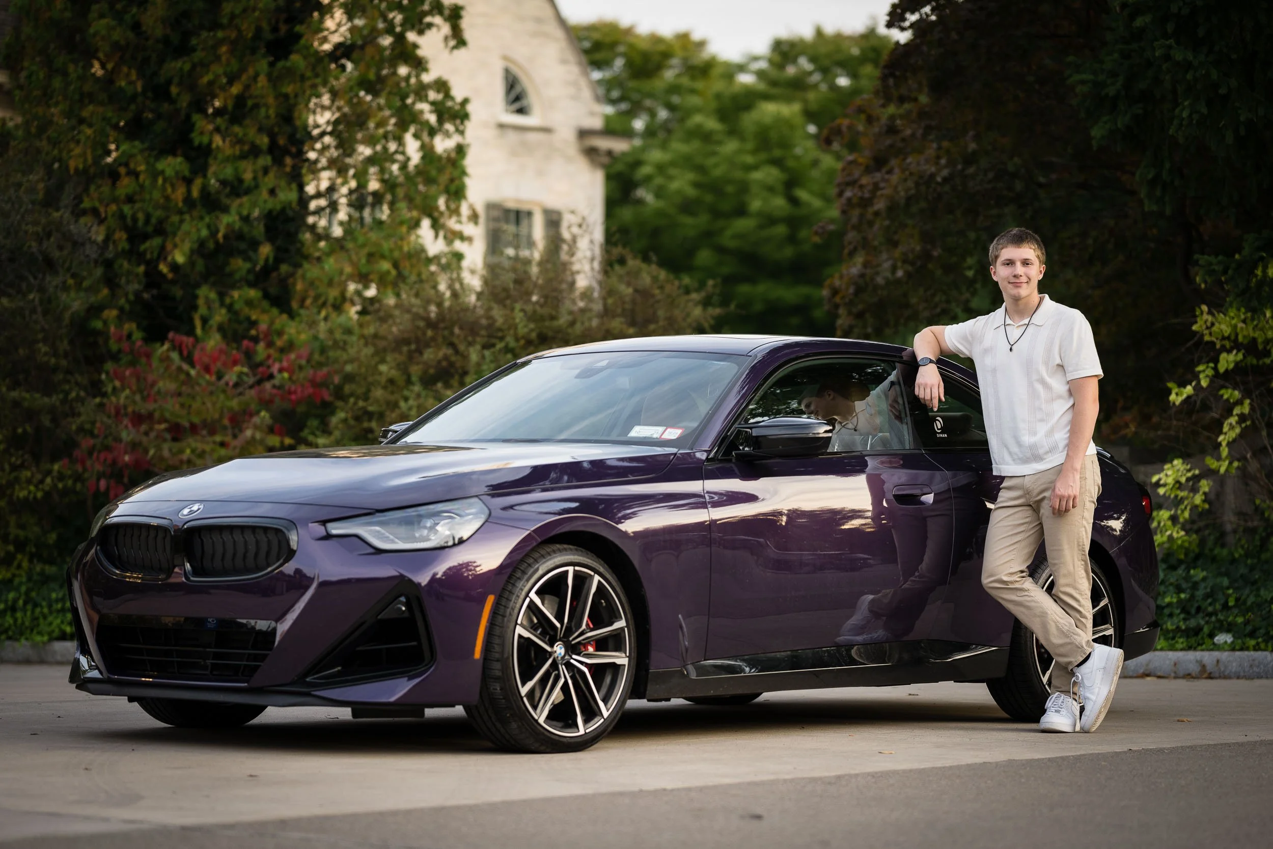 A young man leaning on the roof of a dark purple sports car, standing on a driveway with a background of trees and a stone house. The young man is smiling, wearing a white polo shirt, khaki pants, and white sneakers.