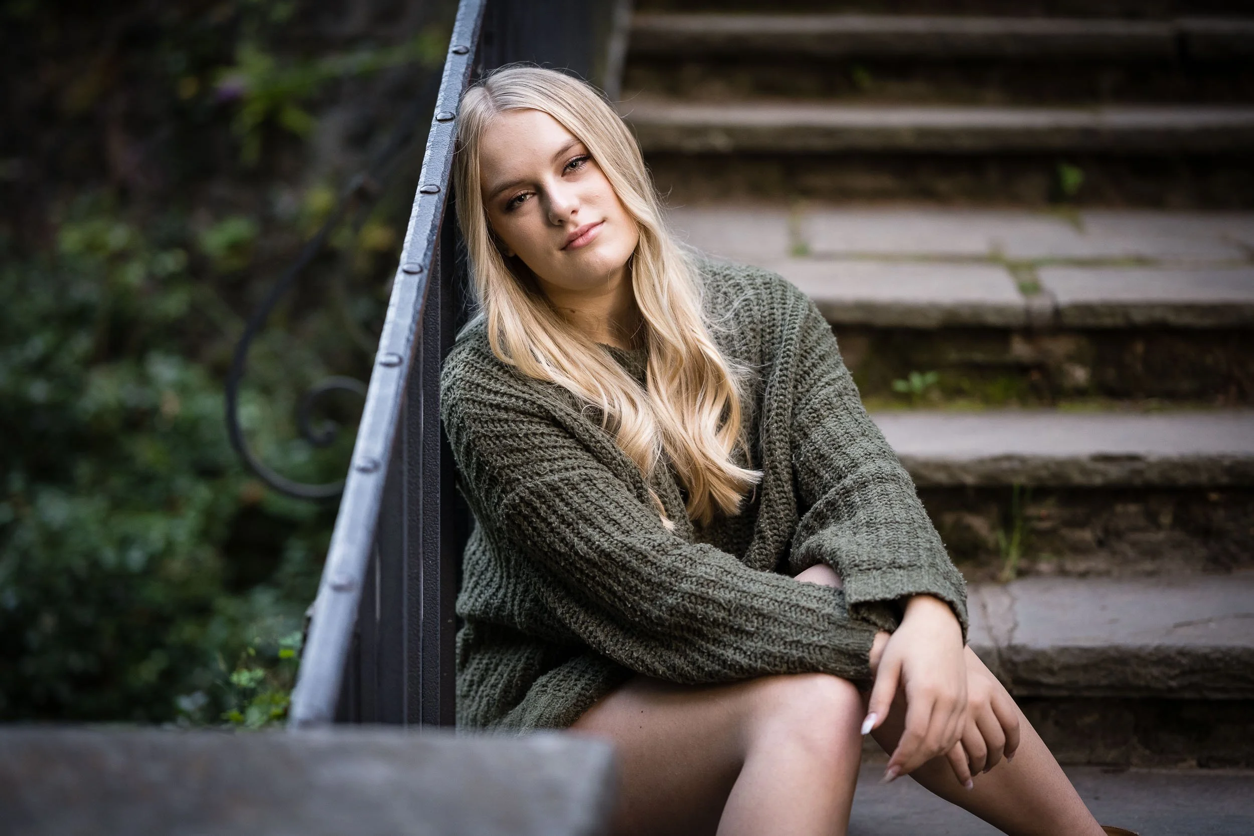 A young woman with long blonde hair sitting on outdoor steps, wearing a green knitted sweater, looking at the camera with a relaxed expression.